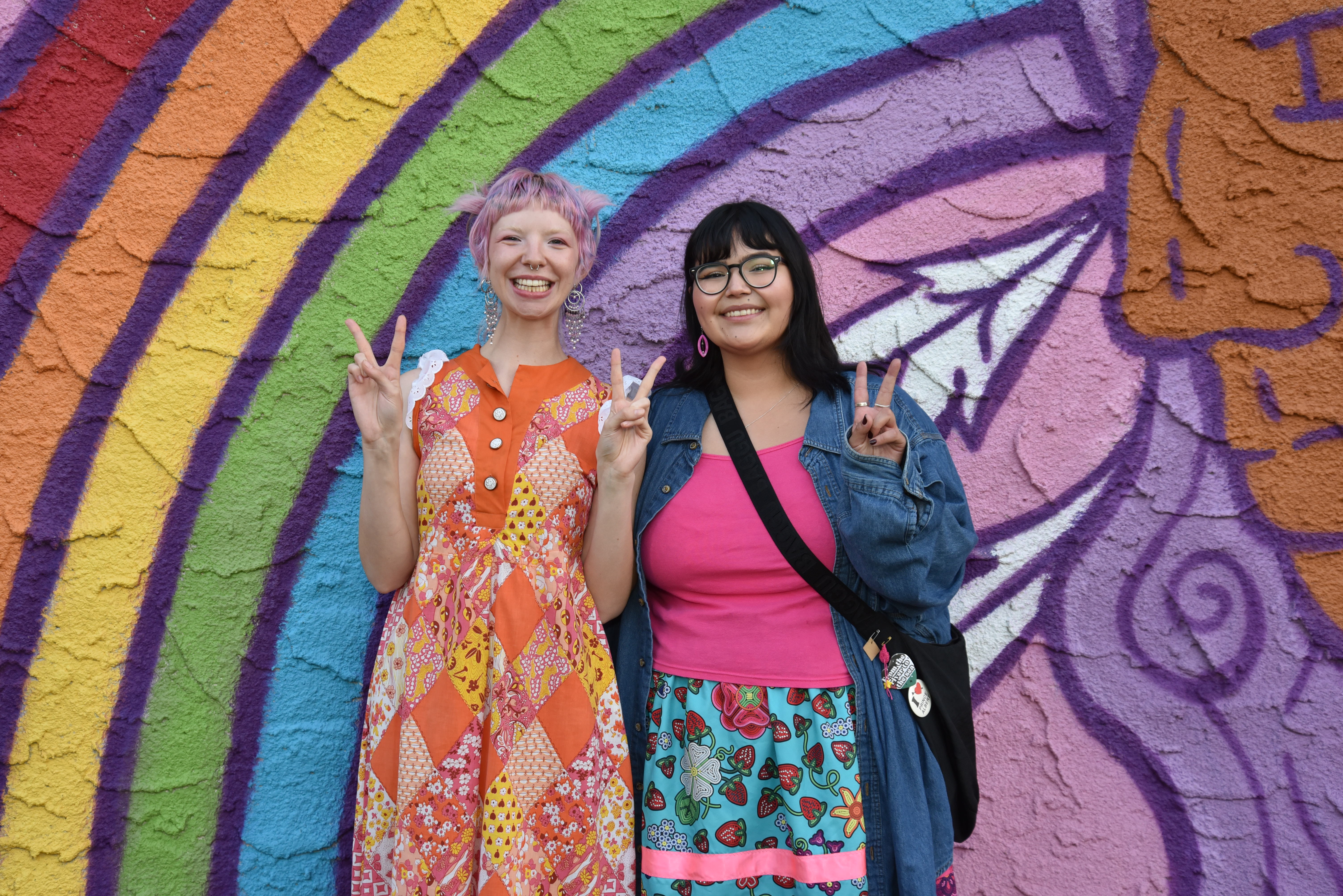 Two ladies dressed colourfully smile and make peace signs with their hands in front of a rainbow mural