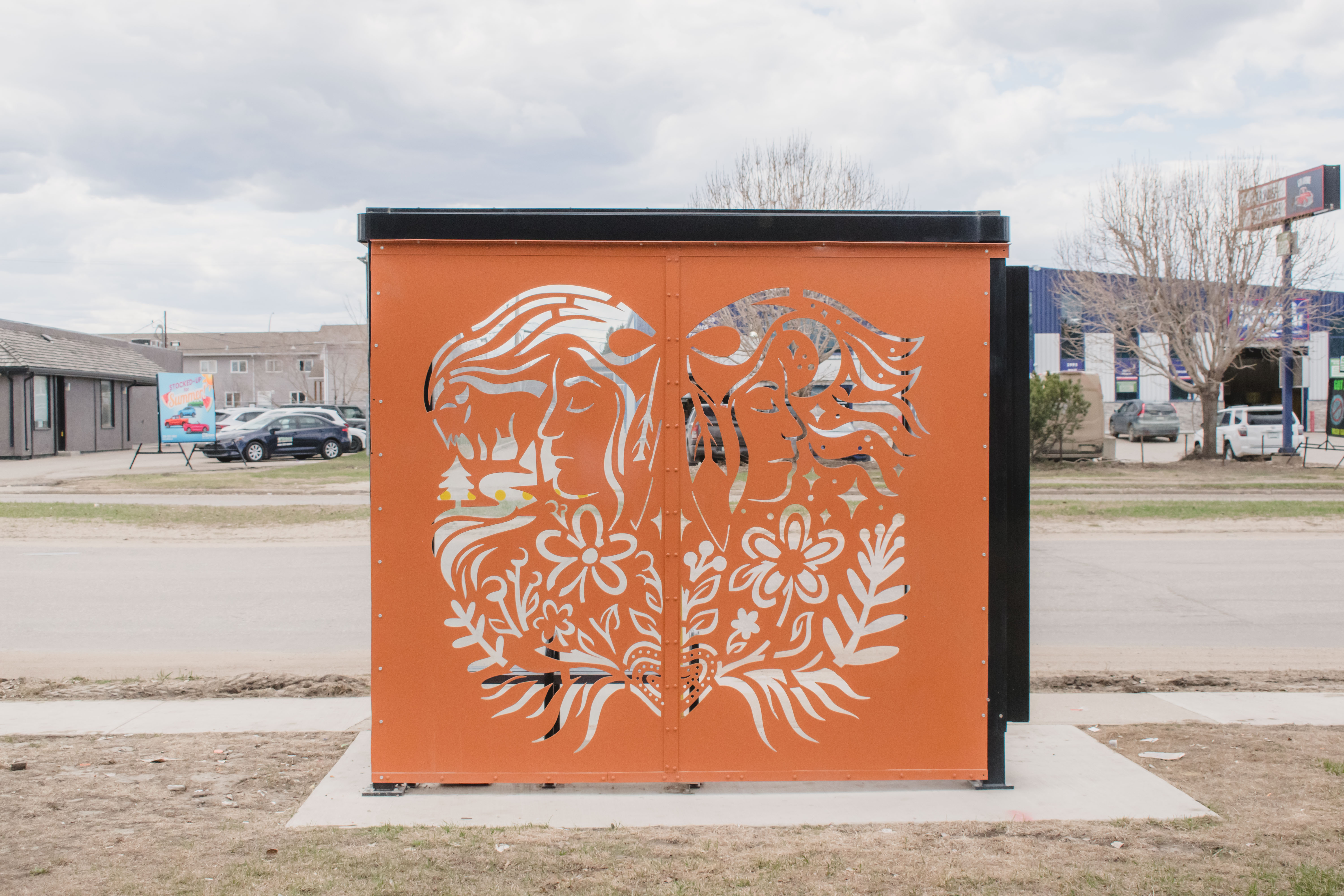 A transit bus shelter with an orange-brown metal design on the back with flowers, leaves, stars, water trees and two side profiles back to back.