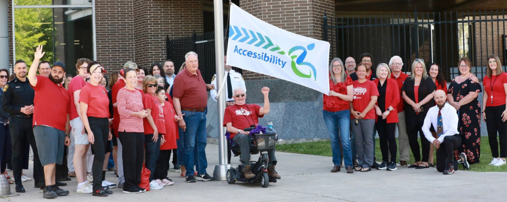 Participants, many wearing red, hold an Accessibility flag by a flag pole at the County of Essex Administration Building.