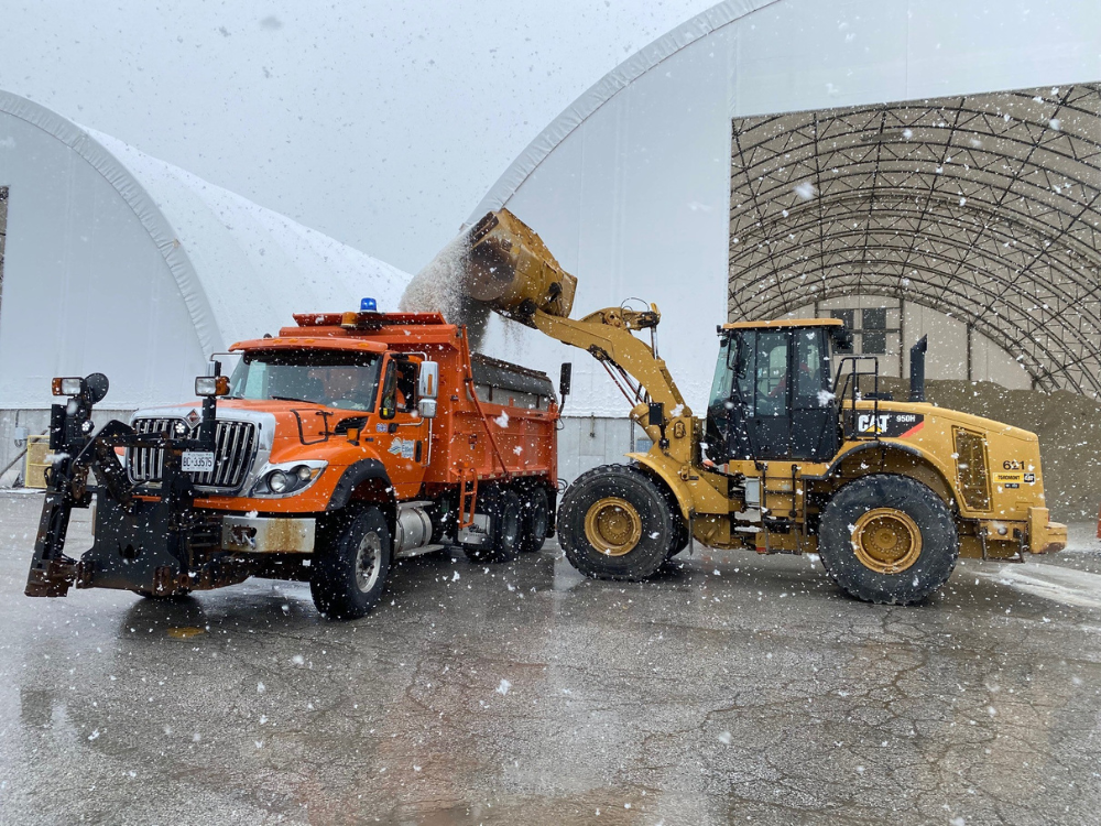 A vehicle loads salt into a salt truck with snow flying.