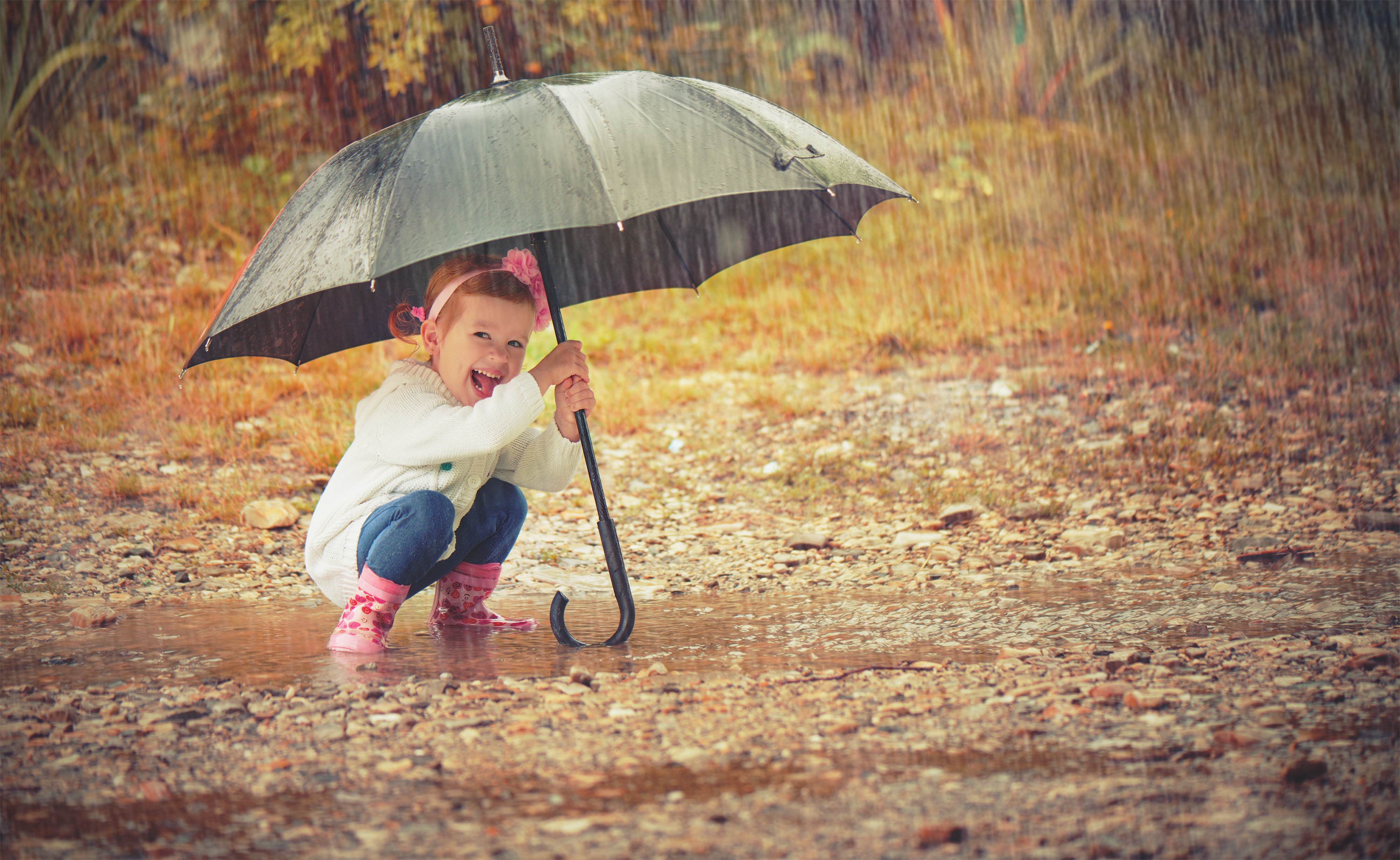 Girl squatting in a puddle, holding an umbrella in the rain with colourful fall leaves on the ground.