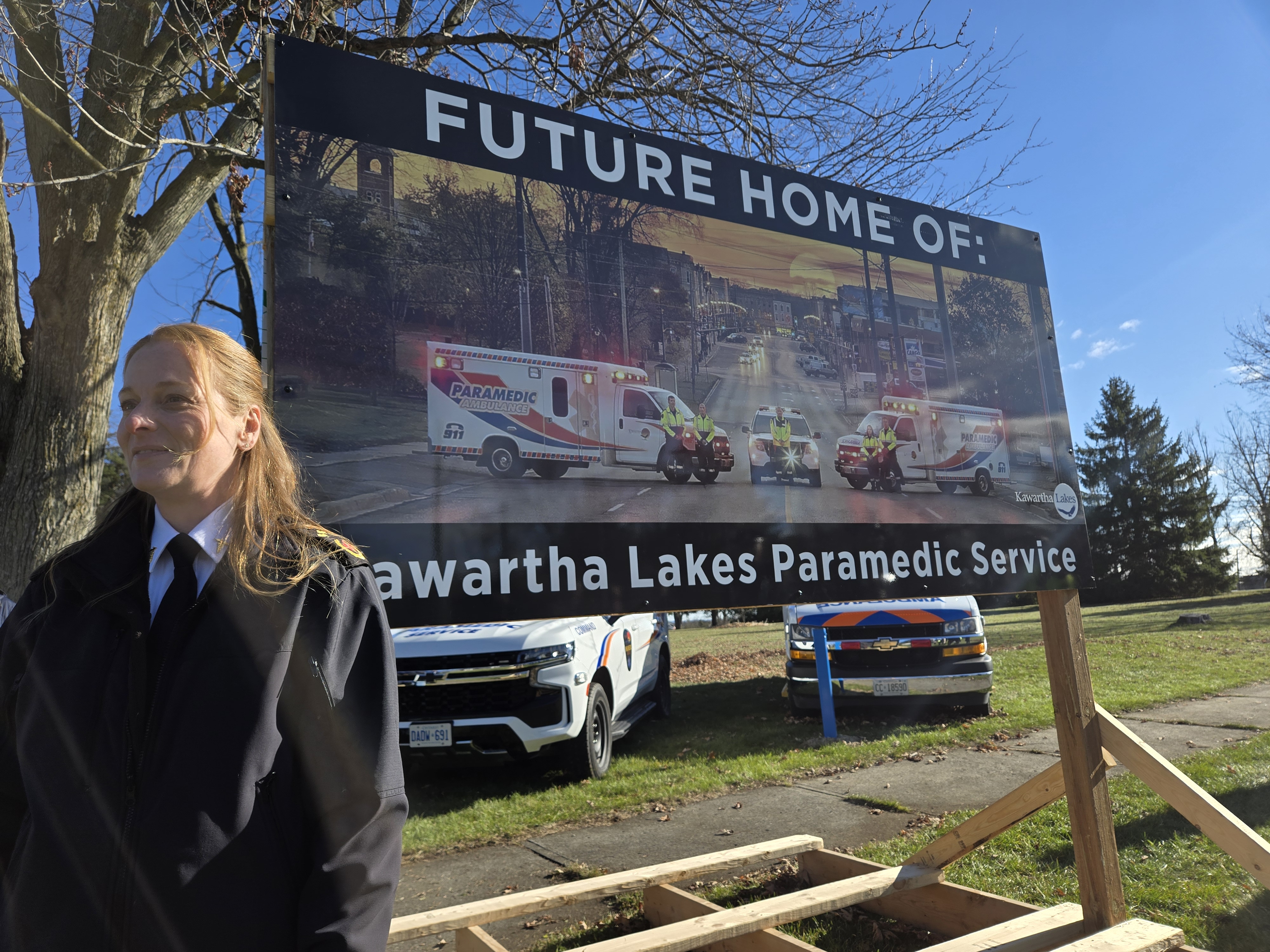 A woman stands outdoors in front of a large sign that says FUTURE HOME OF: Kawartha Lakes Paramedic Service