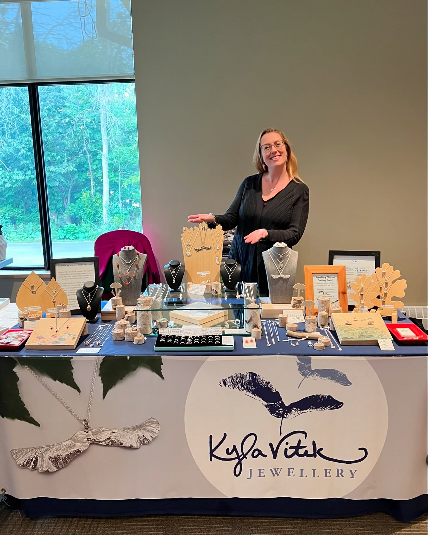 Woman in black clothing smiling and standing behind her table display of handmade jewellery.