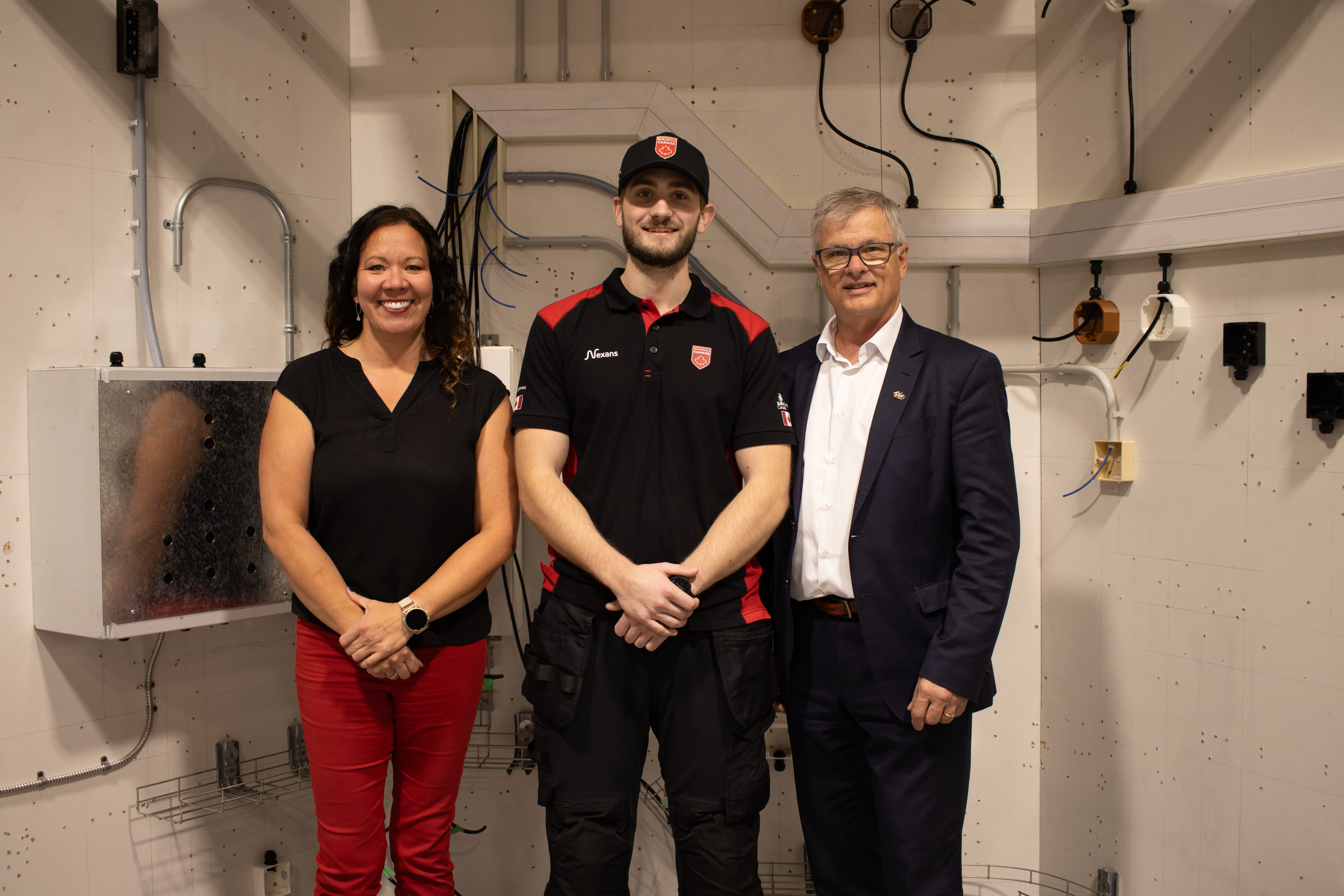 Three people pose together in an electrical training lab with wiring and electrical equipment in the background.
