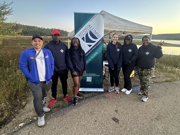 Keyano College Alumni Association members pose together at an outdoor event beside an alumni banner and canopy.
