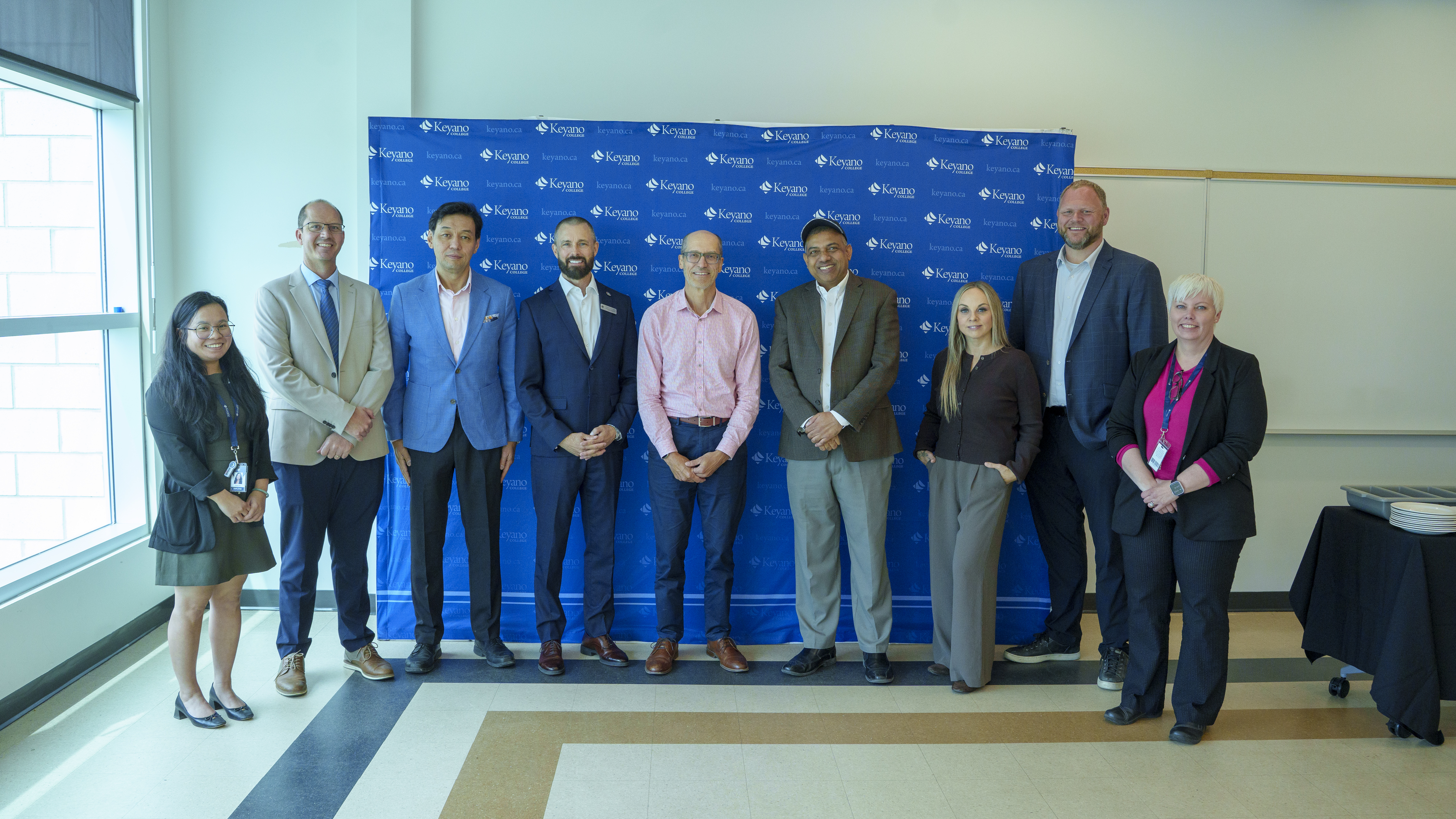 A group of nine individuals pose for a photo in front of a Keyano College backdrop during a campus event. Participants are dressed in business and professional attire and stand together in a classroom or meeting space.