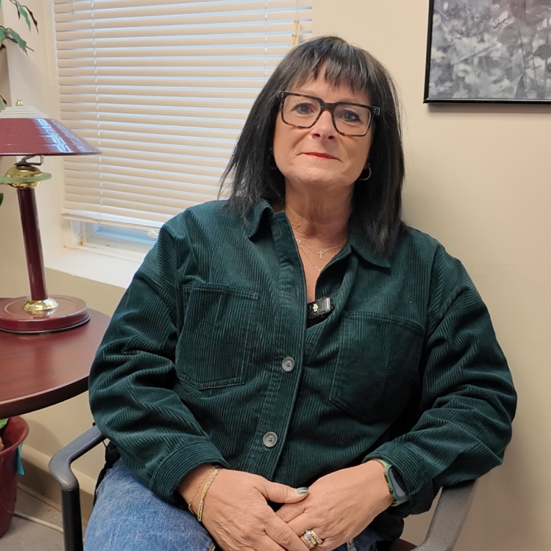 A woman with shoulder-length dark hair and glasses sits in an office chair beside a desk lamp and a window with blinds. She wears a green corduroy jacket and a microphone clipped to her collar.