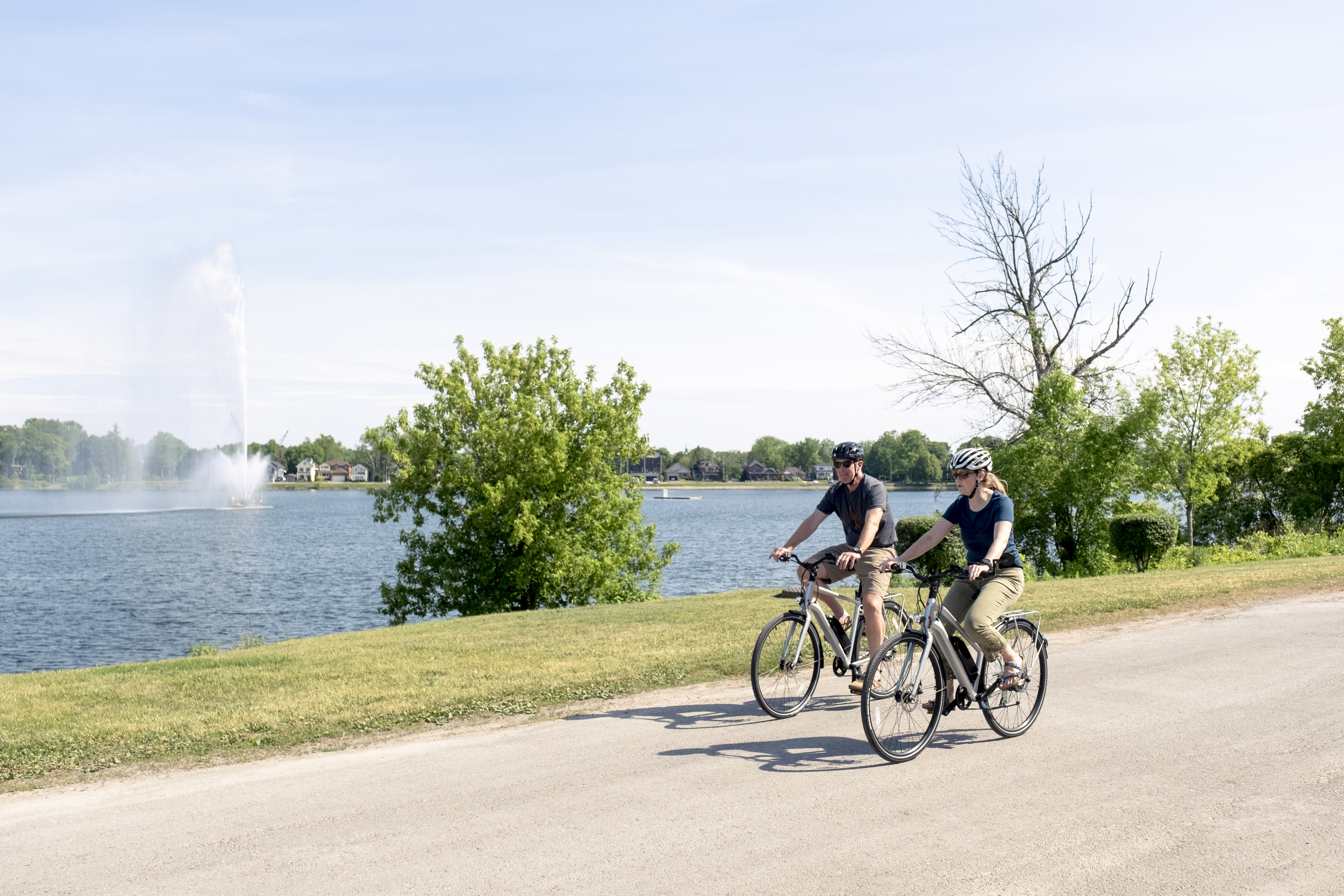 A couple cycling on a roadway; Peterborough's Little Lake is in the background with the Centennial Fountain running - sky is blue, early 