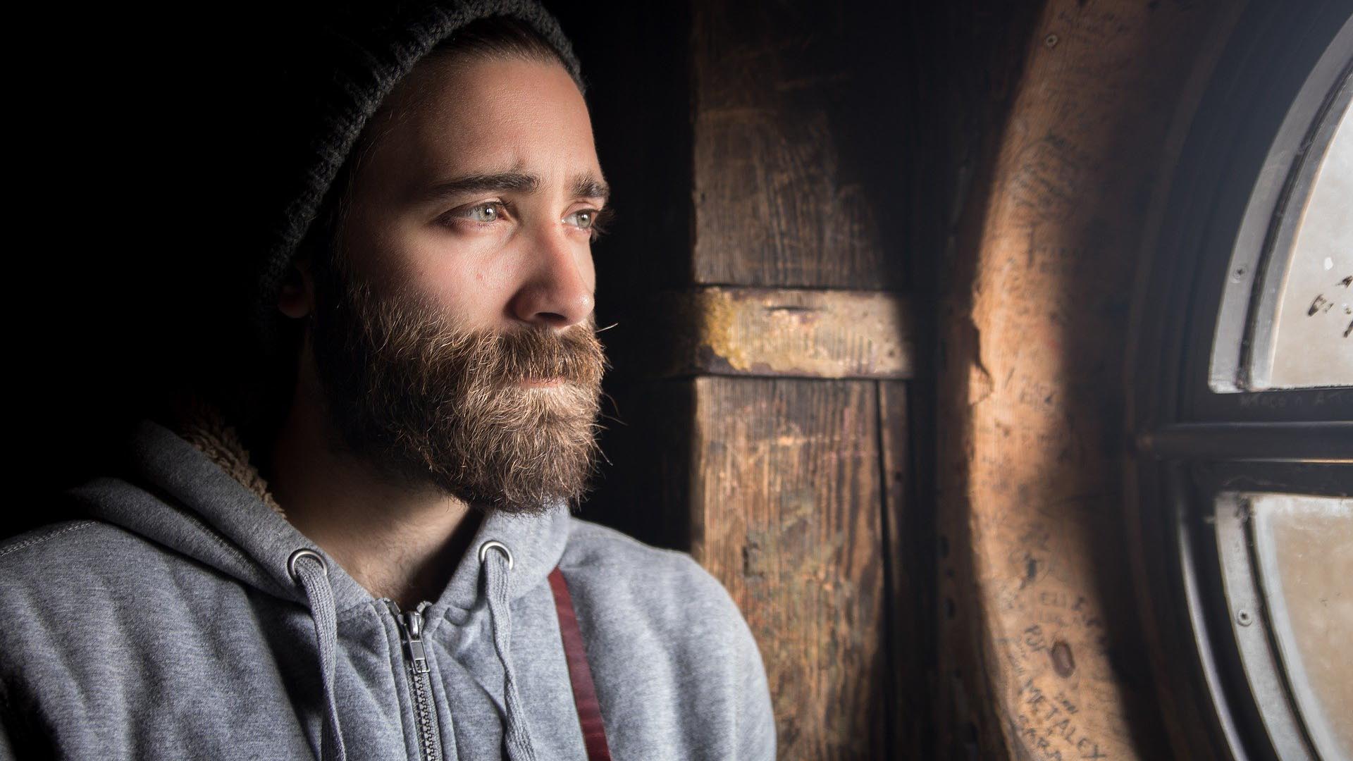 Man with a beard, wearing a beanie, looking out a window