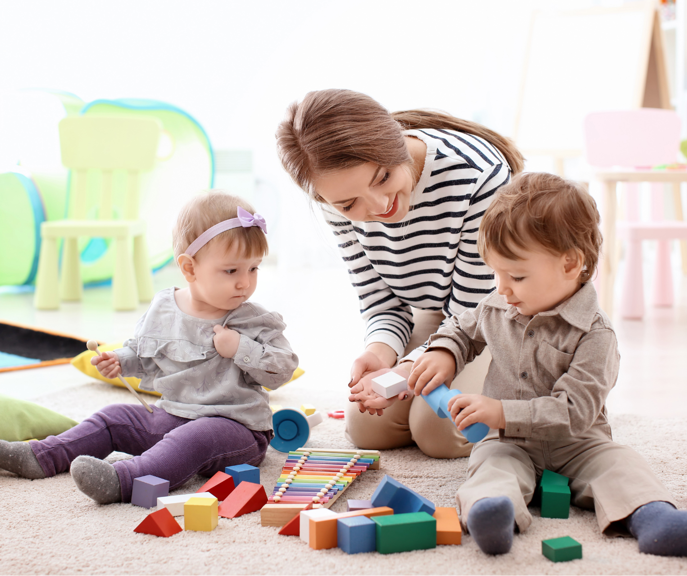 A woman sits with two babies playing with wooden blocks on the floor