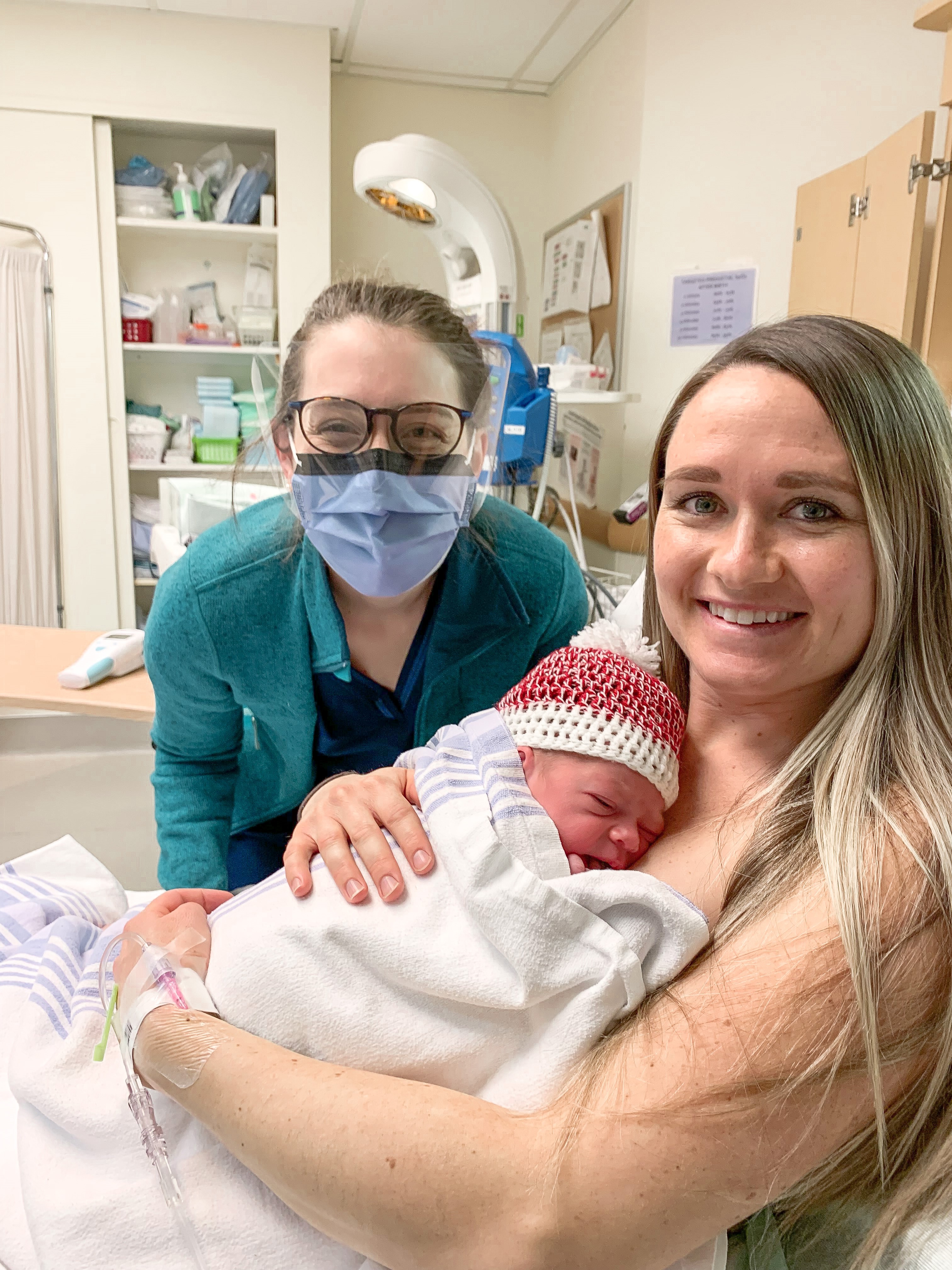 A doctor crouches beside a woman in a hospital bed, holding a newborn, in a hospital room