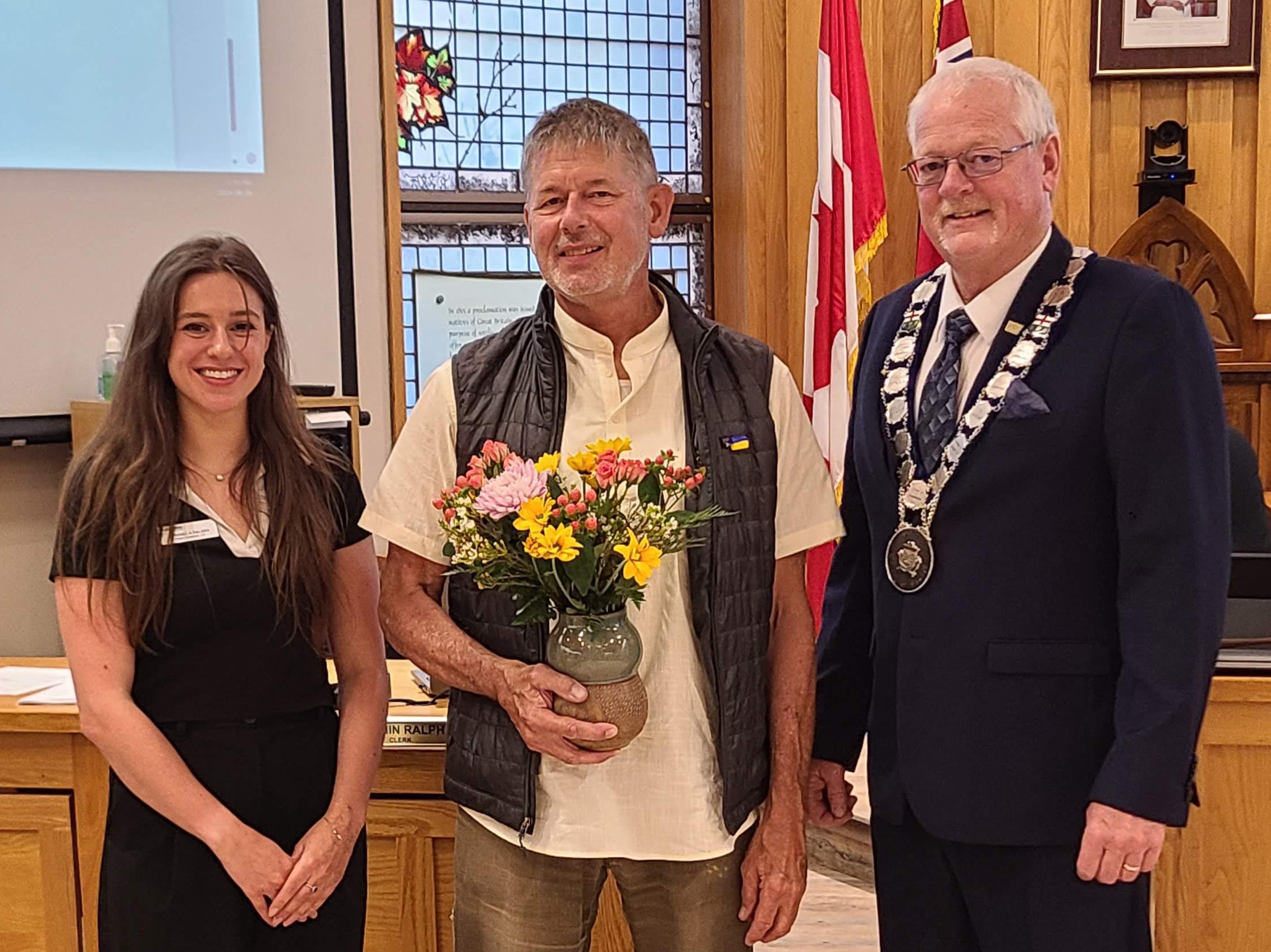 Three people stand together for a photo. In the middle, a man holds a vase of flowers.