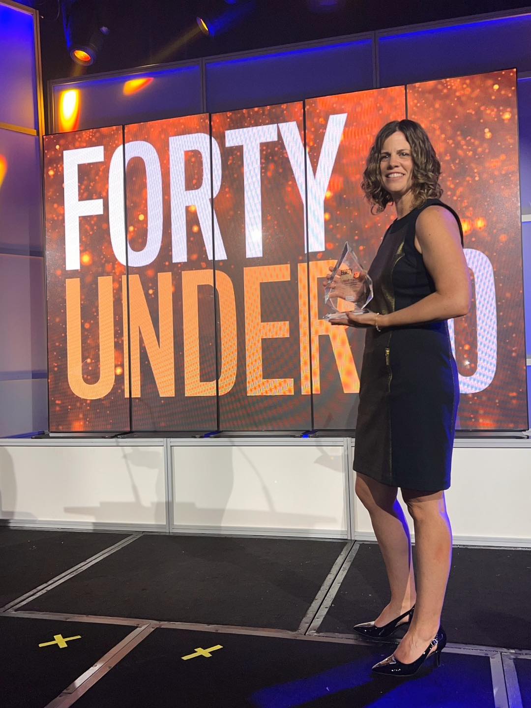 Woman with brown shoulder length curly hair in a black dress, holds a clear glass award in front of a video screen that reads 'Top Forty Under Forty'