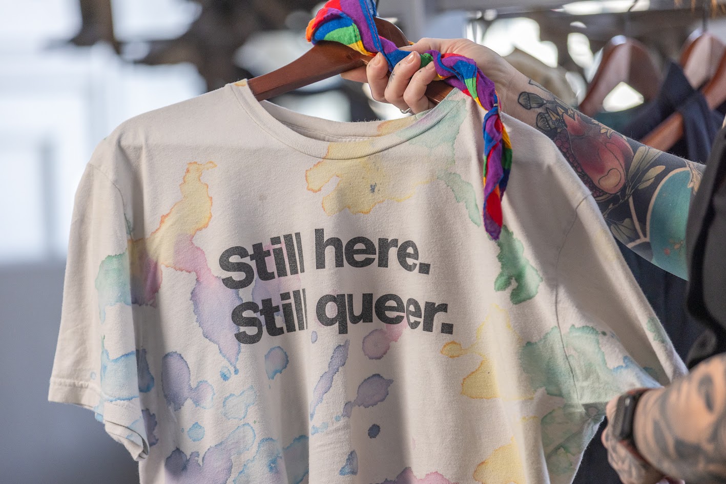 A person holds up a tie-dyed shirt with the phrase 'Still here. Still queer' in black lettering.