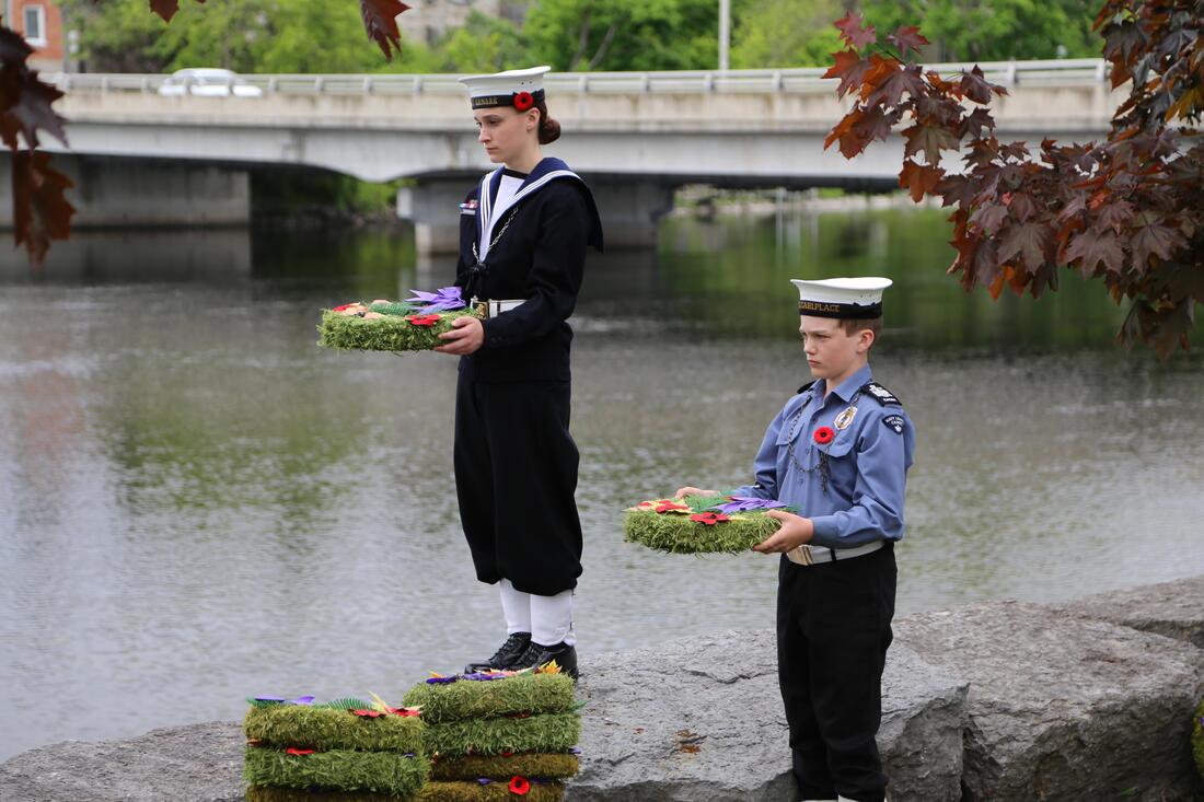 Two cadets in full uniform hold wreaths by the water's edge