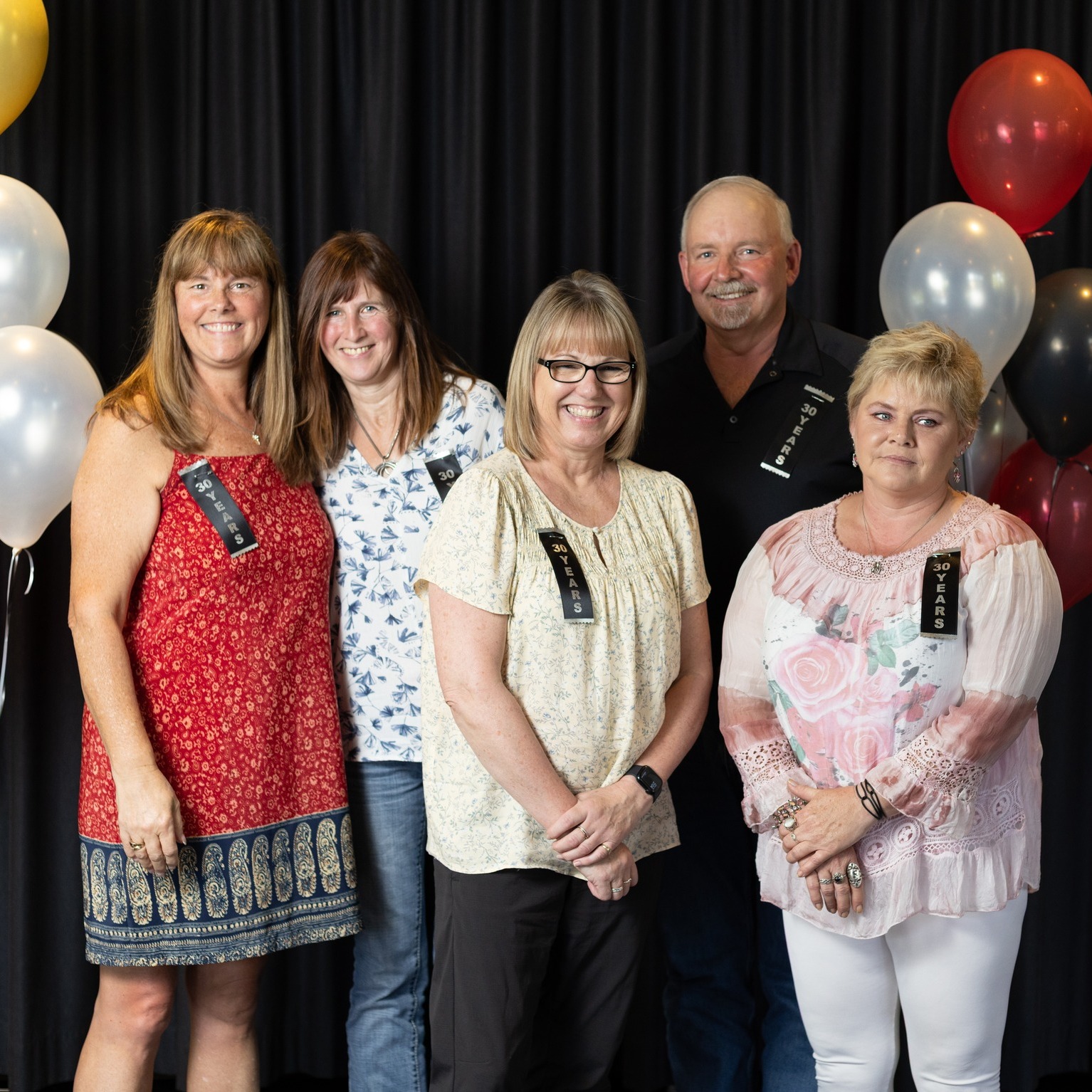 A group of people smile for a photo against a black backdrop with balloons