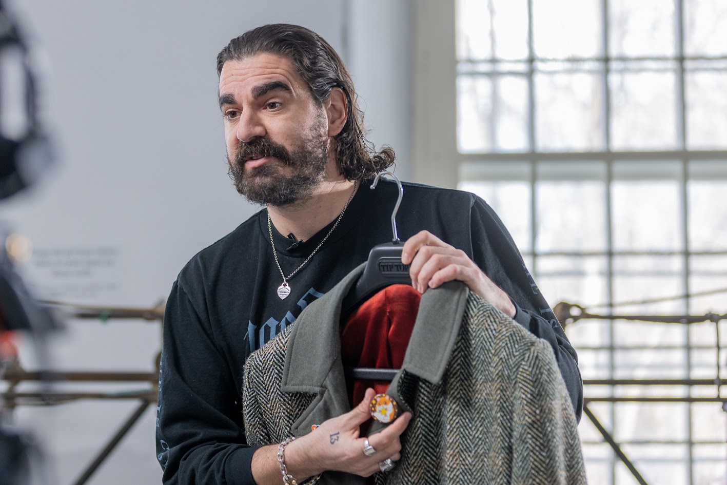 A man with long, dark hair and a beard, wearing a black shirt, displays a grey peacoat on a hanger