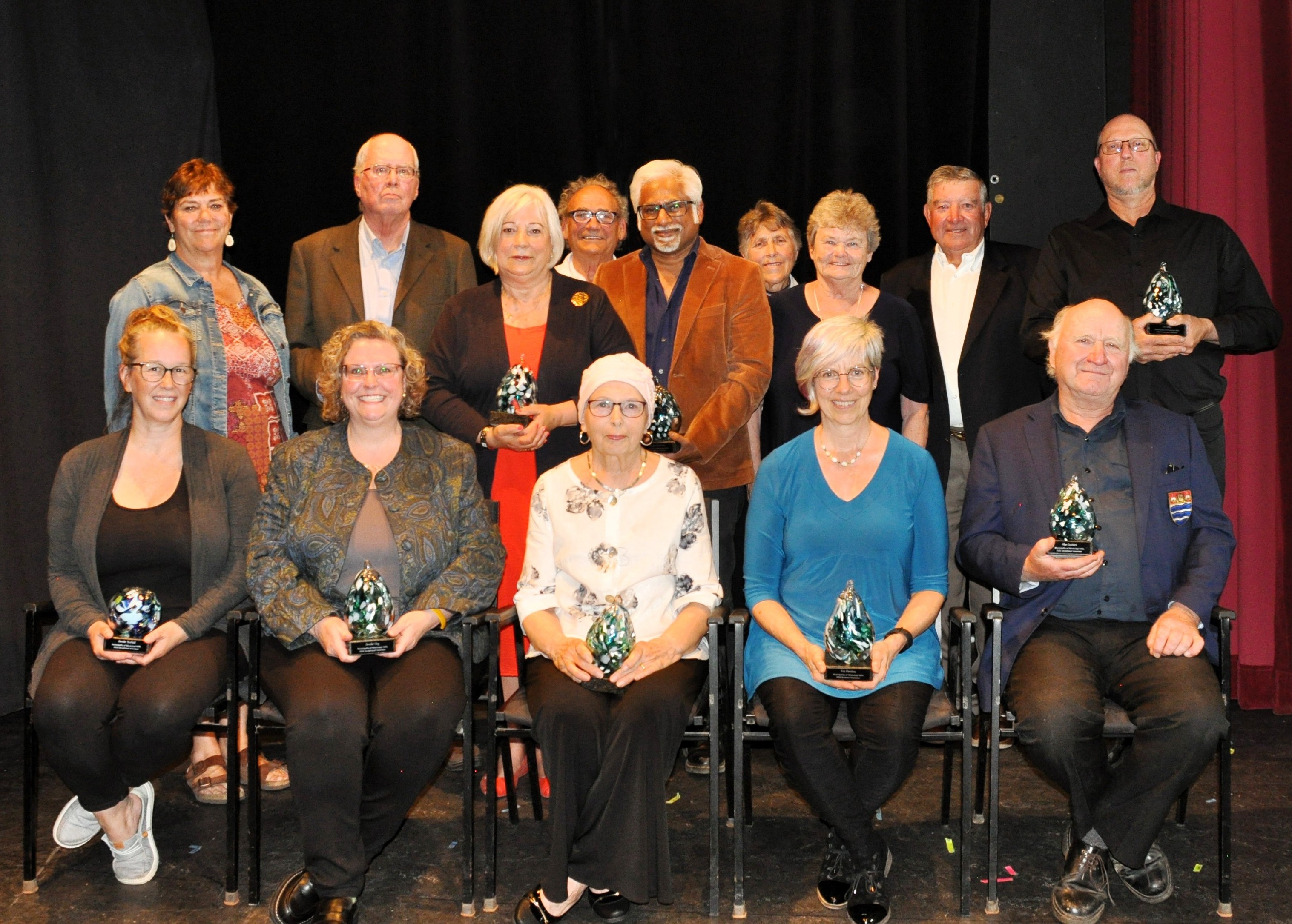A group of people, standing in the back row and seated in the front row, hold awards on a stage with a black curtain.