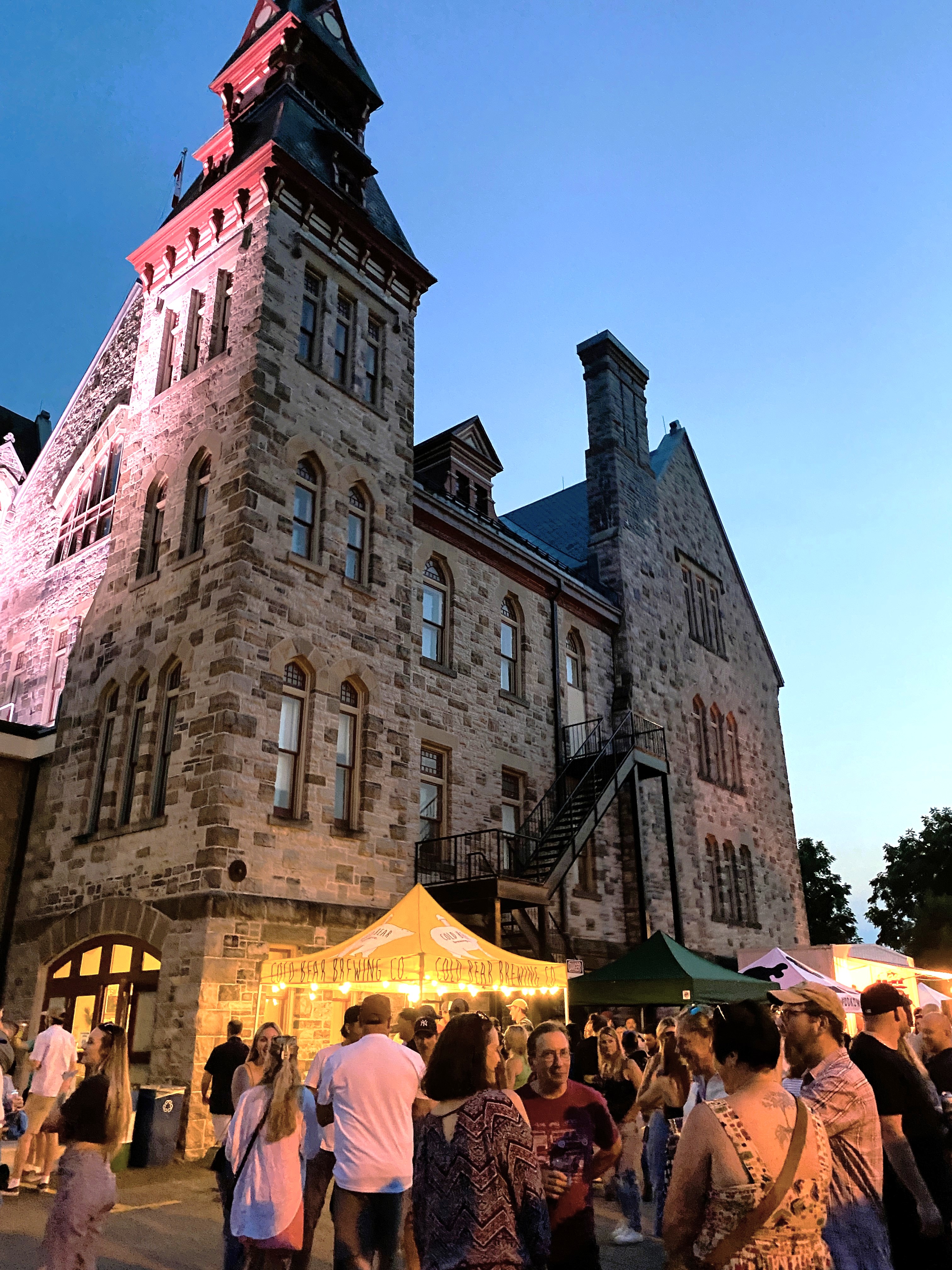 Tents are set up with groups of people standing in front of a large stone building at dusk