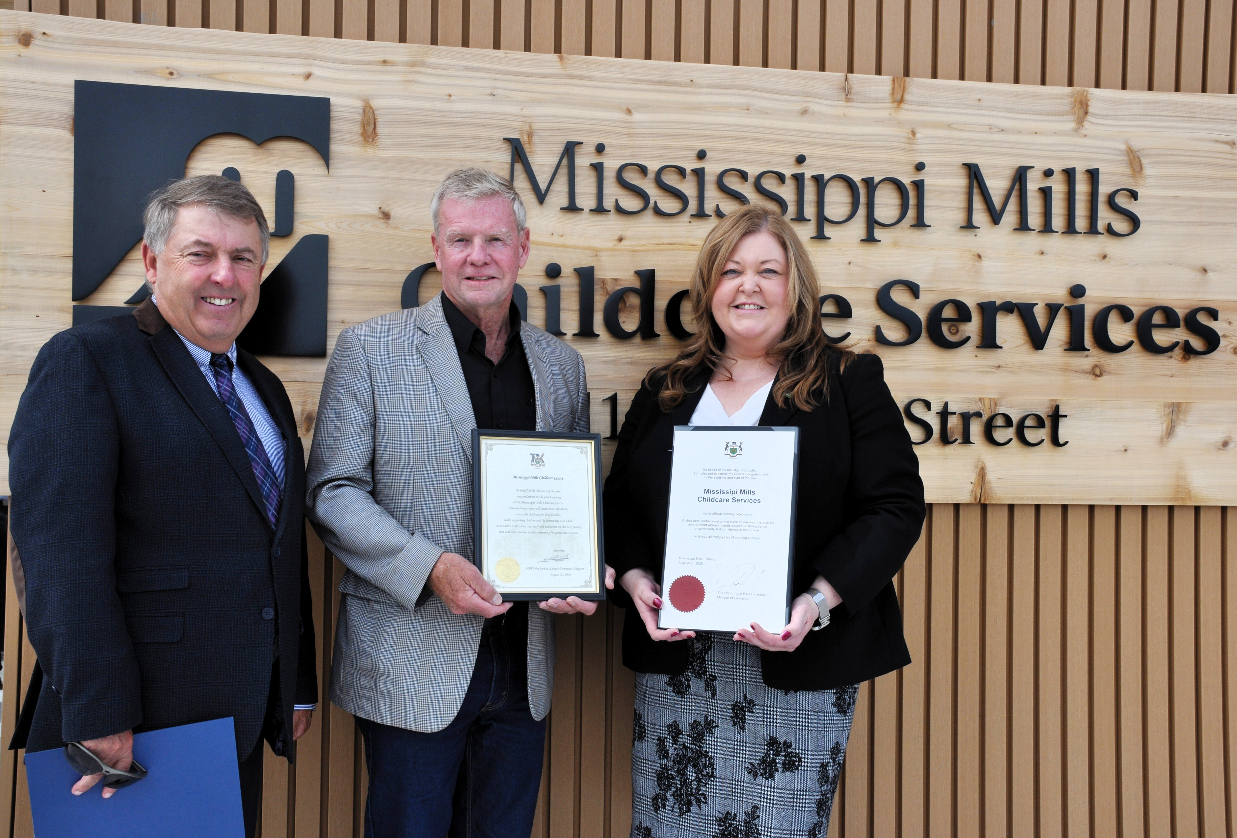 Two men and a woman stand in front of a wooden sign reading 'Mississippi Mills Childcare Services'. The man in the middle is holding a certificate and the woman on the right is also holding a certificate.
