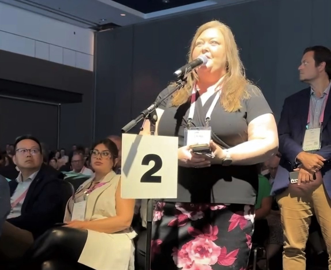 Woman stands in a conference room at a microphone, surrounded by people
