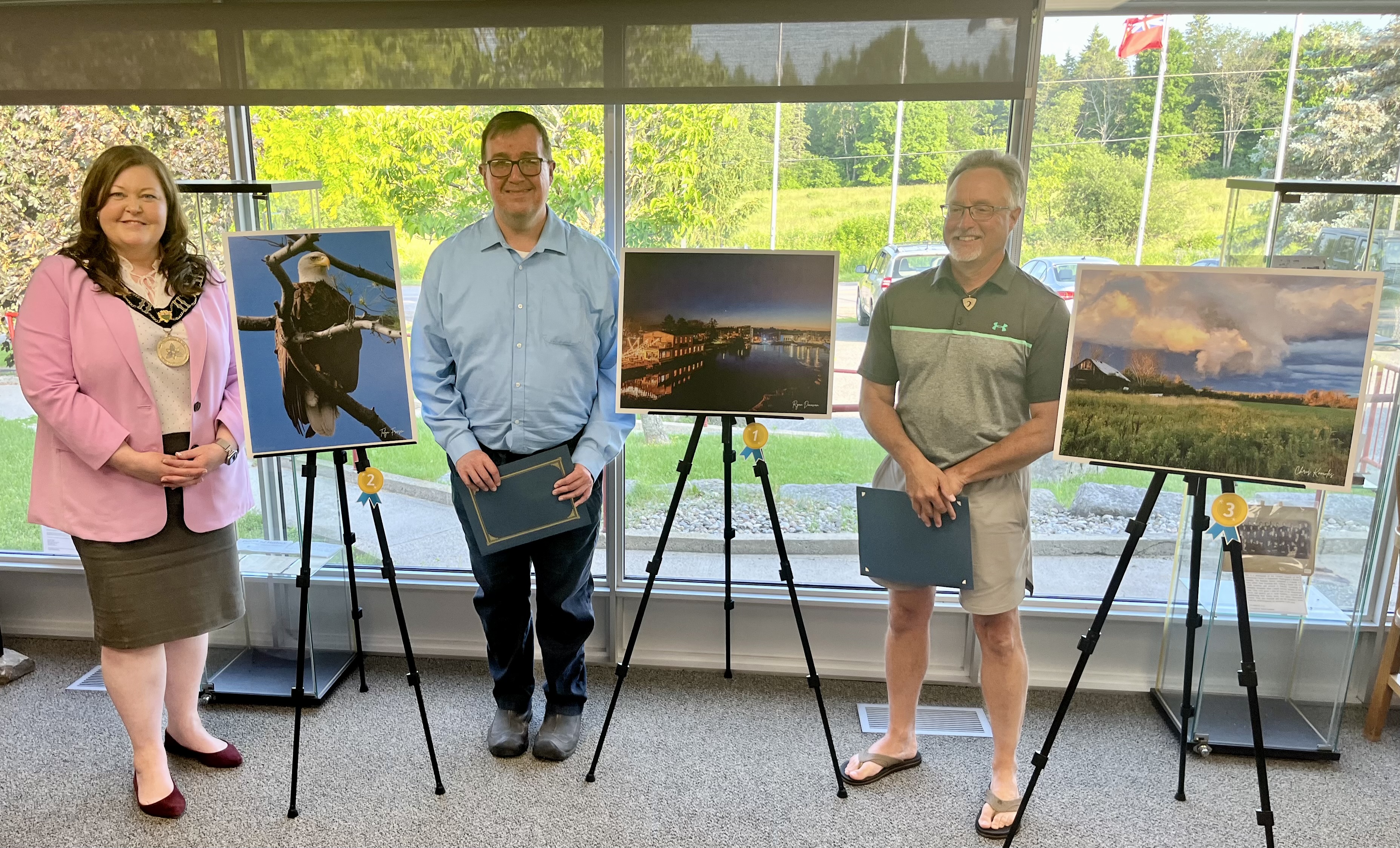 A woman and two men stand in a room next to large photographs on easels.