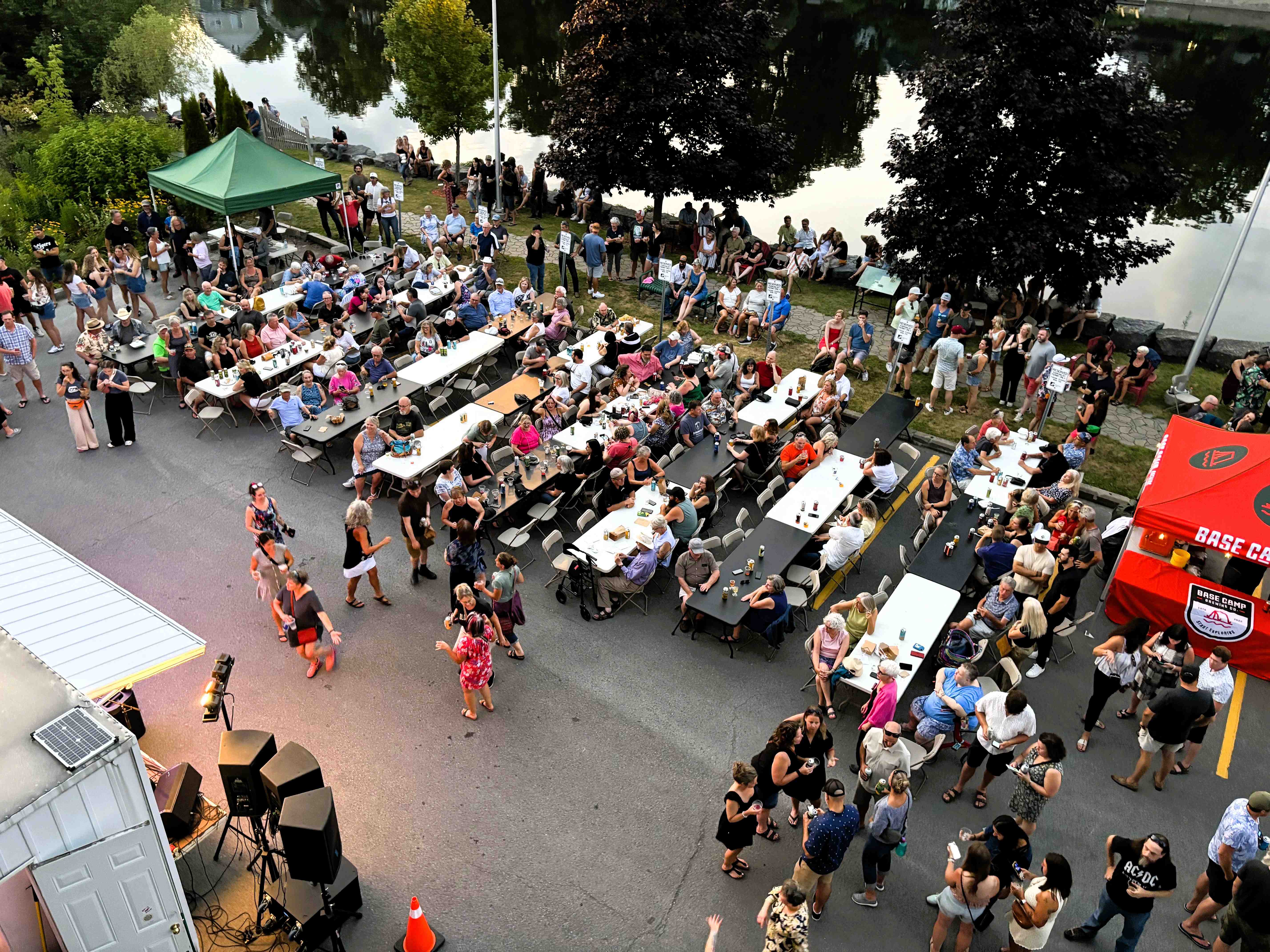 From above, people sit at rows of tables in a paved parking area next to a river. Some people are up dancing near a stage.