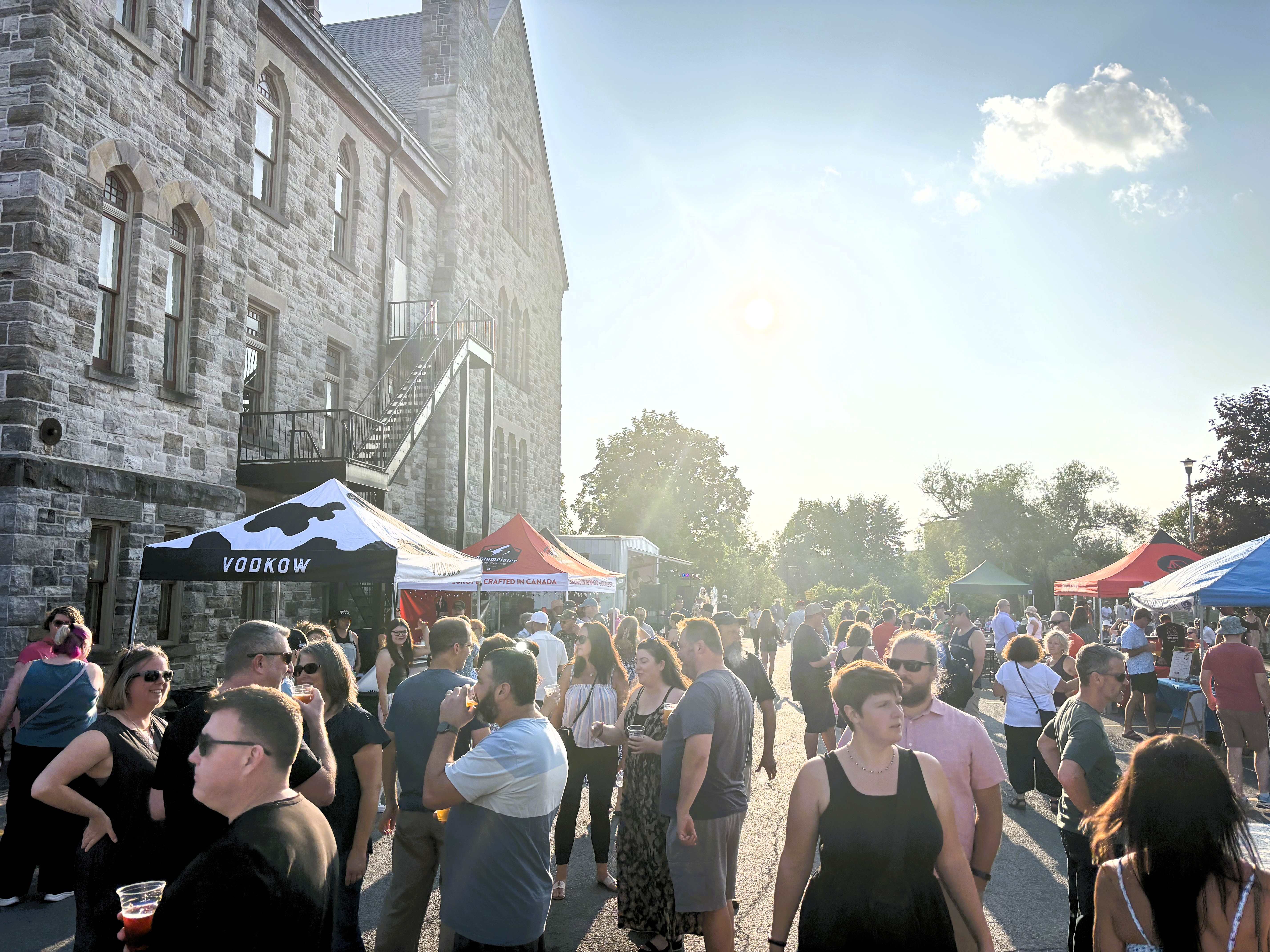 People gather under sunny skies in a parking area adjacent to an old stone building.