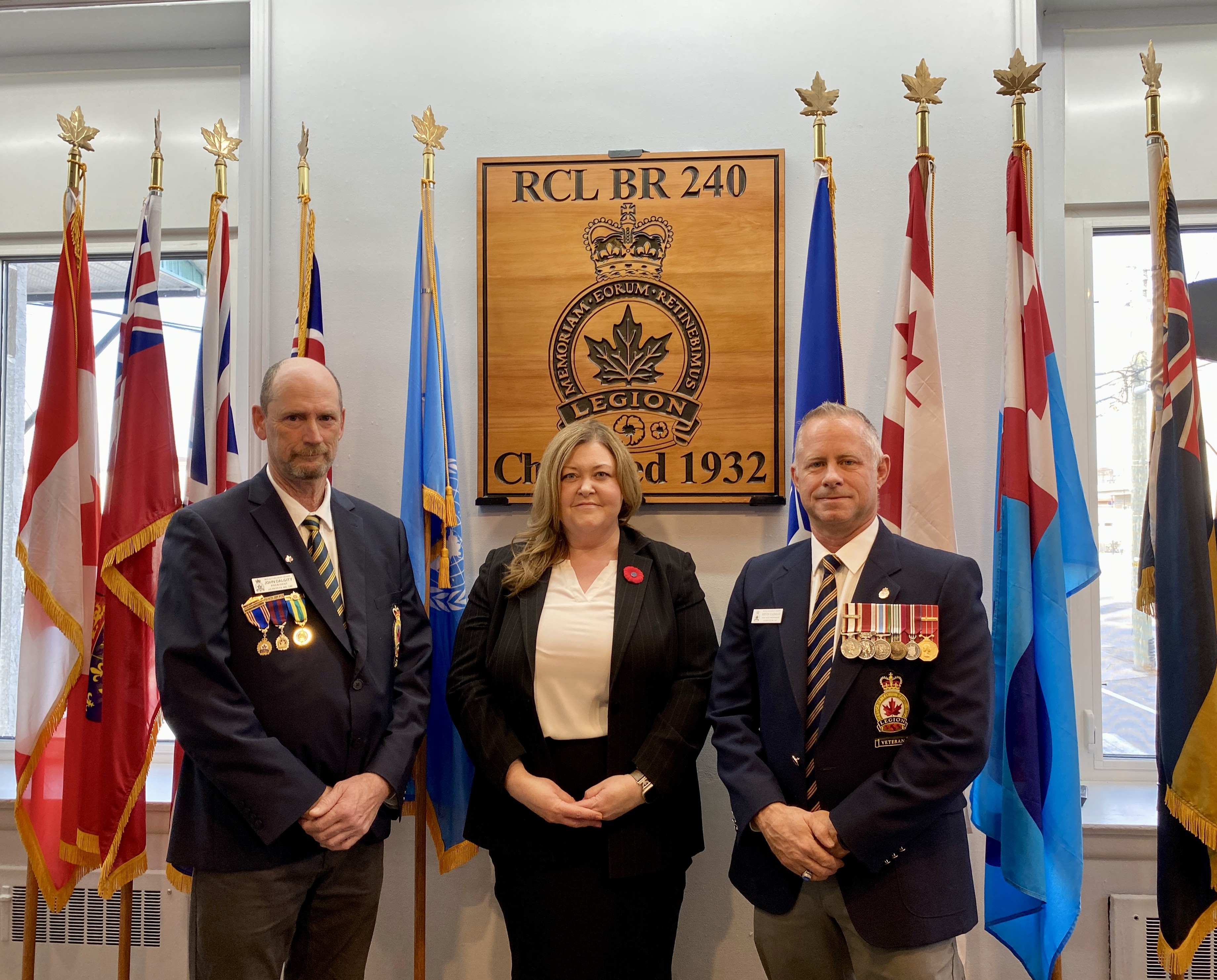Three people stand in front of a group of brightly coloured flags. A wooden plaque hangs on the wall between them reading 'RCL BR 240.'