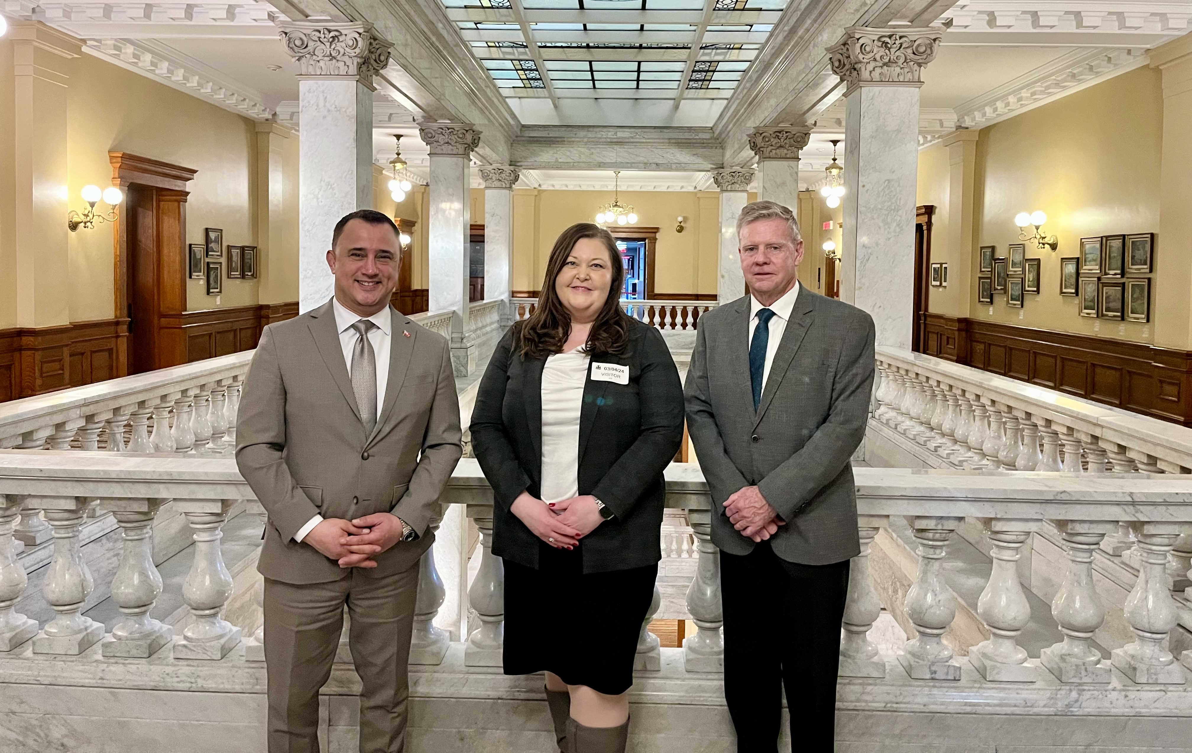 Three people (two men and one woman) stand in front of a marble railing in a regal building