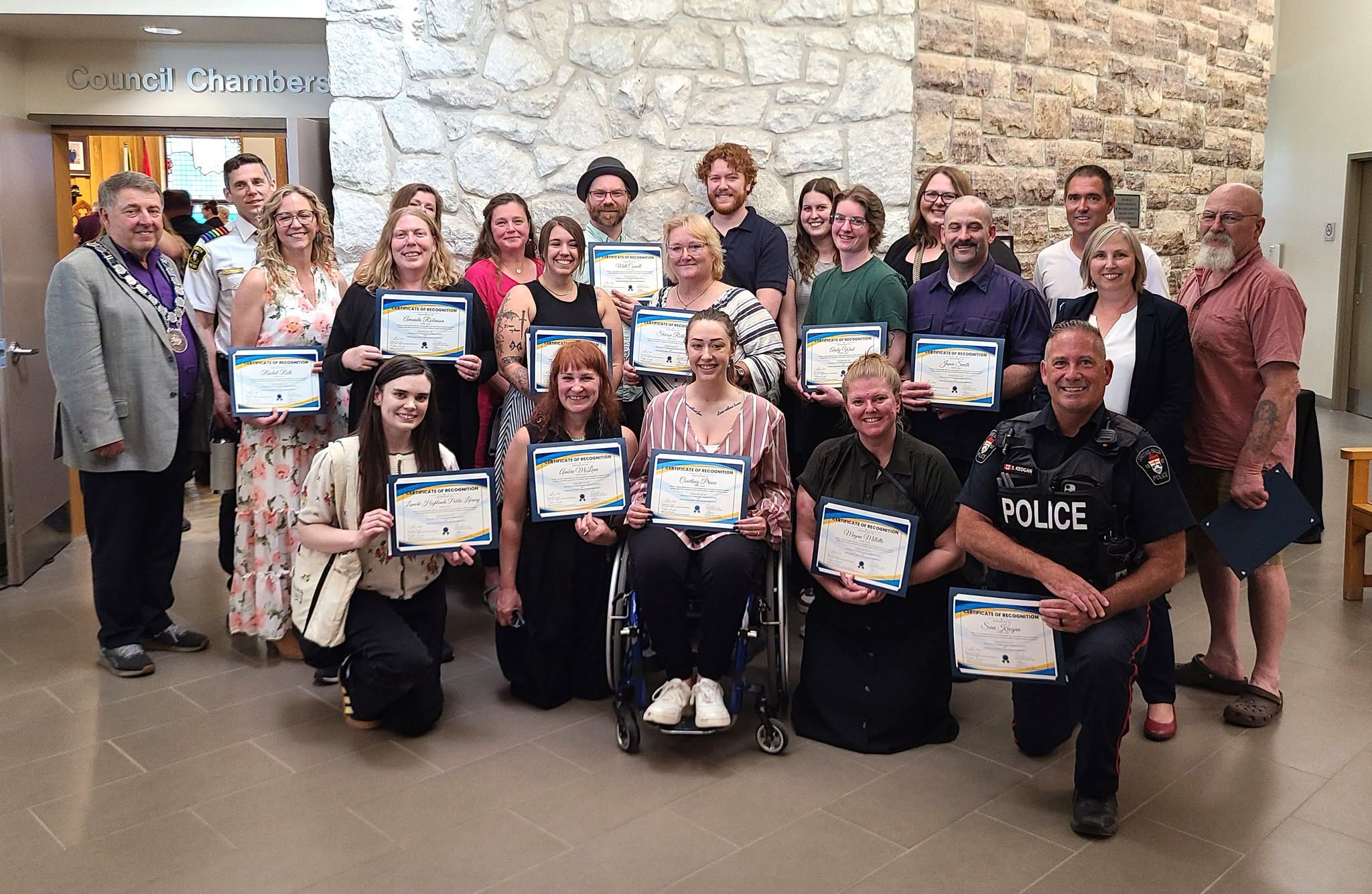 A group of people hold certificates and stand against a stone wall outside a room labelled 'Council Chambers.'