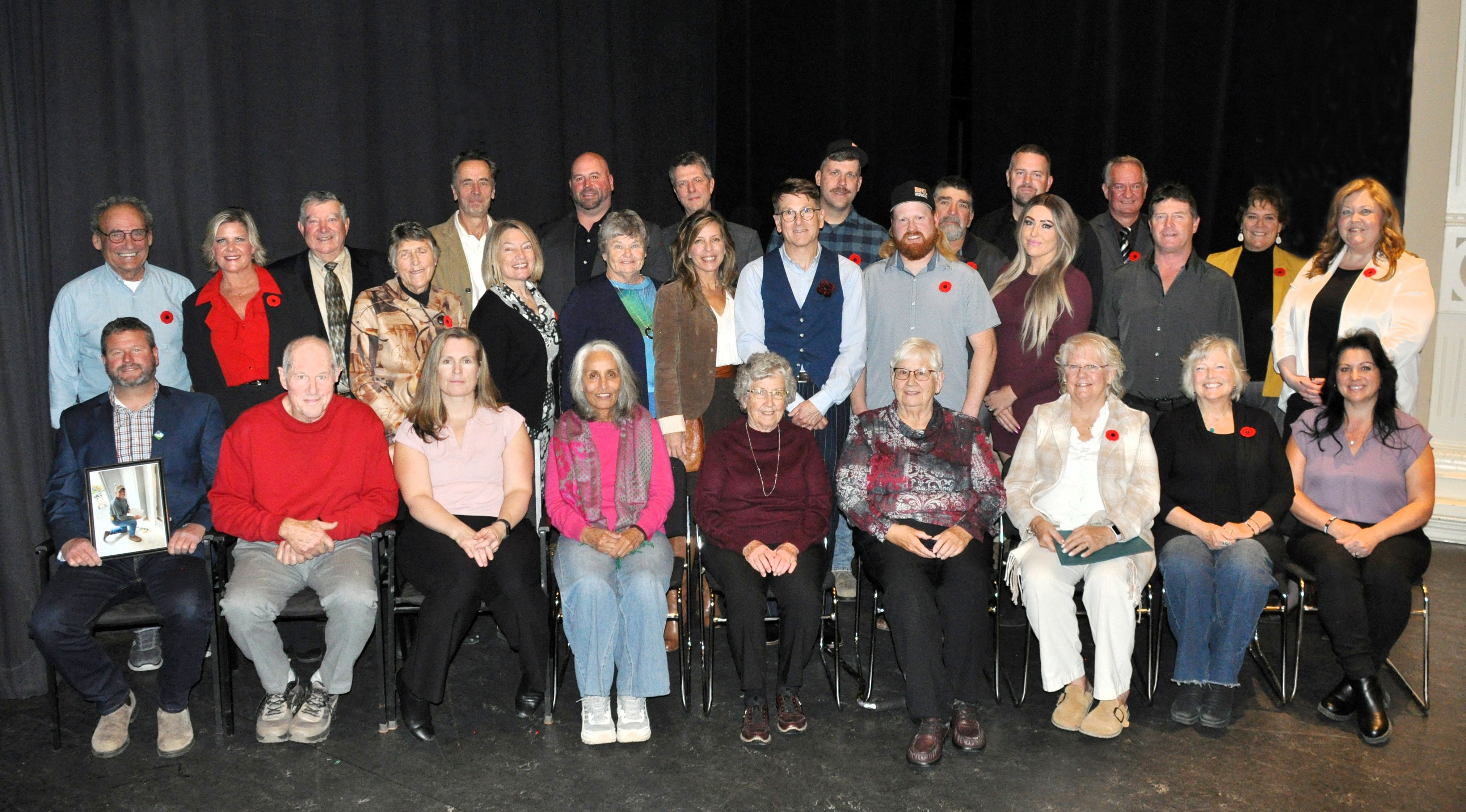 Large group of people pose for a photo on a stage against a black curtain. Back row is standing, front row is seated in chairs.