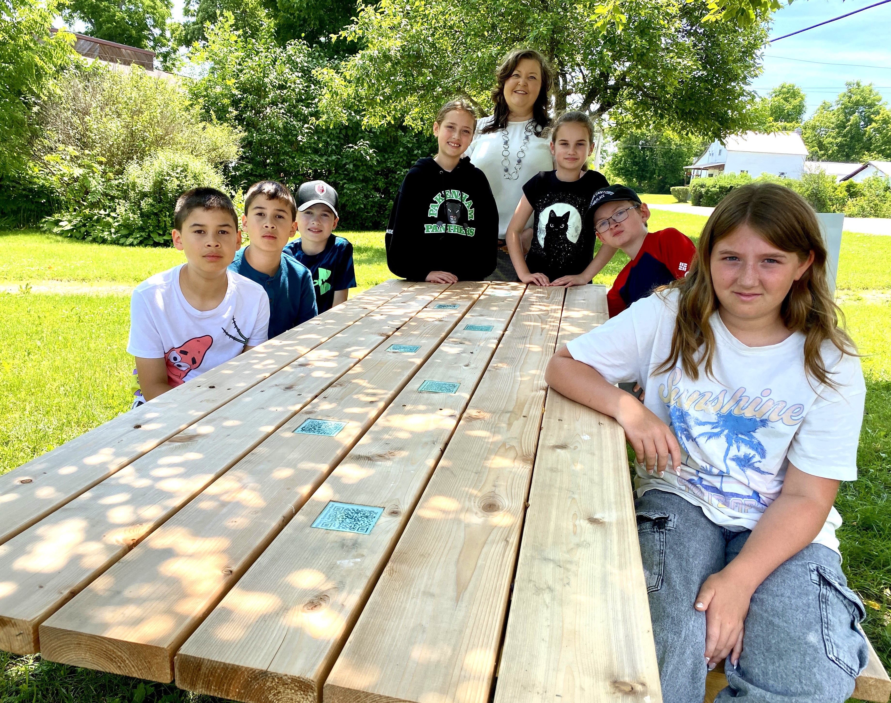 Students and adult woman stand and sit around a wooden picnic table under the shade of a tree