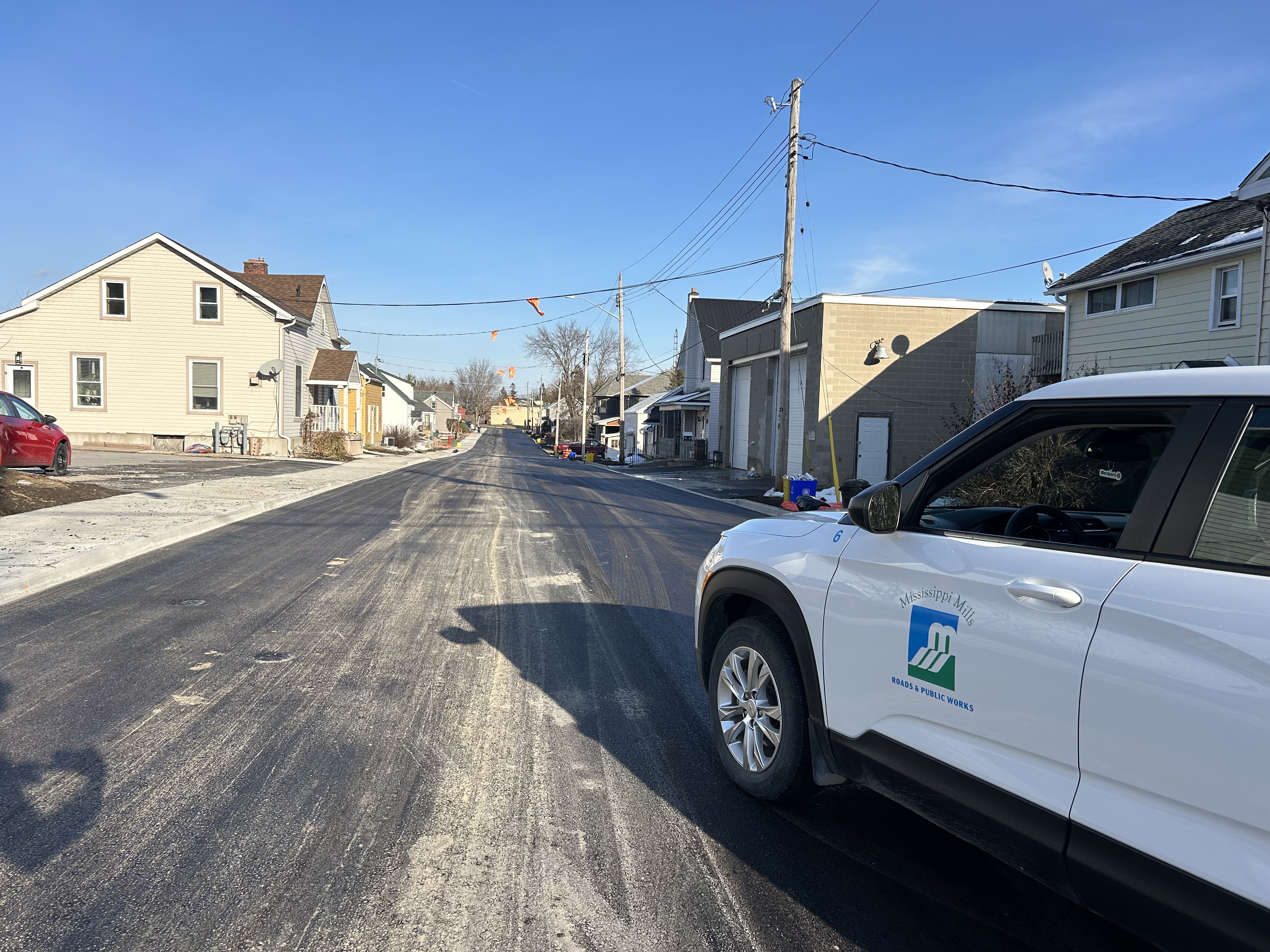 Houses stand in the background of a newly paved street. In the foreground is a white vehicle with the words 'Mississippi Mills Roads and Public Works'