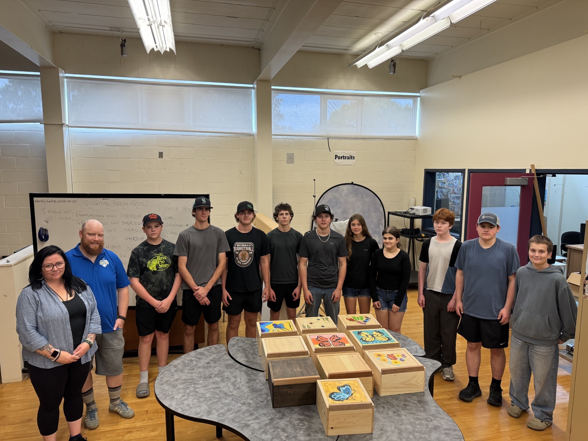 A group of teachers and students stand in a classroom behind a table that holds wooden boxes with decorative designs on the top.