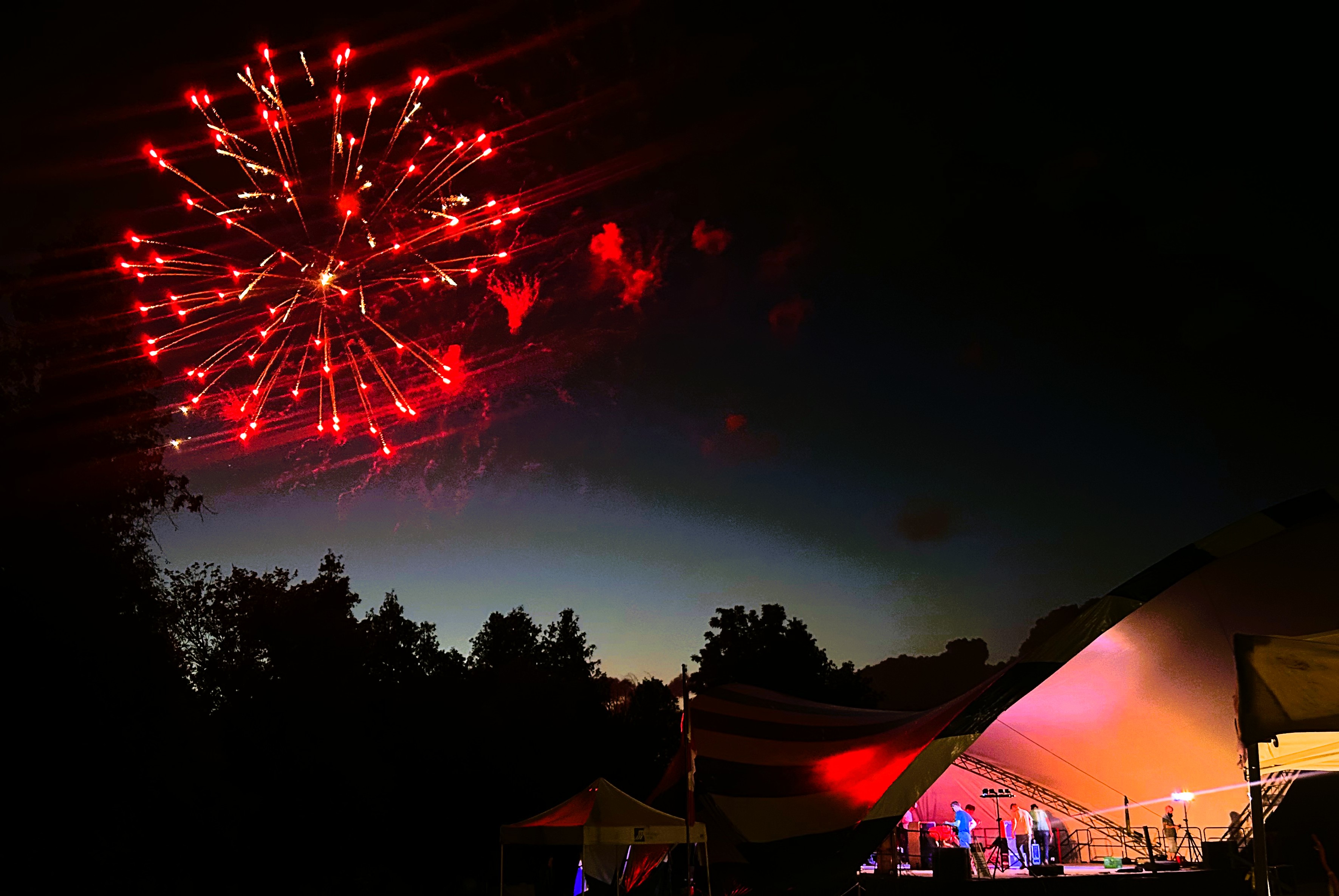 A brillant red fireworks display explodes across the night sky. To the right, musicians perform onstage under a bandshell.