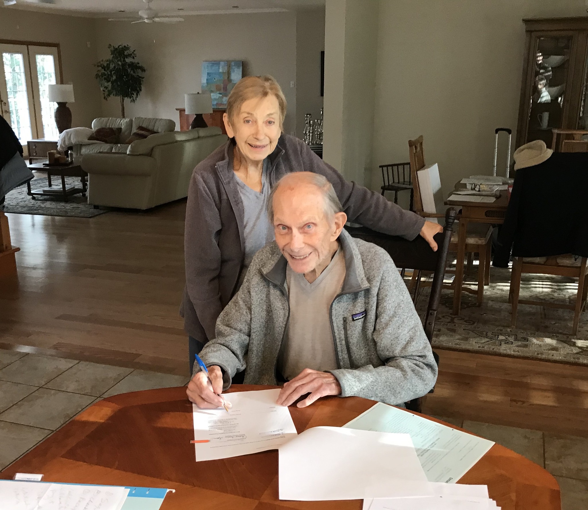 Man sits at table signing a document with woman standing behind him