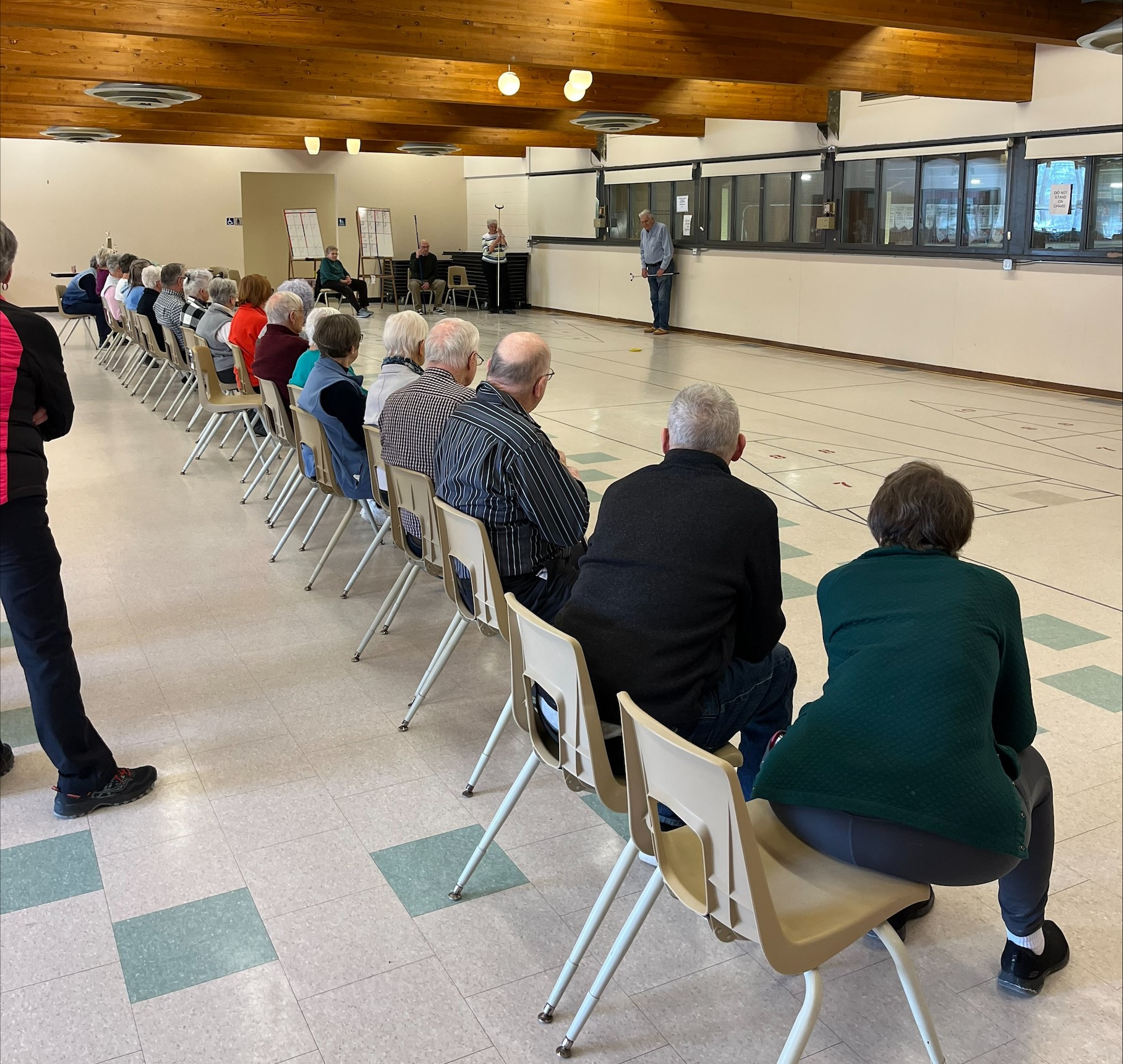 People seated in a row of chairs watching a shuffleboard tournament