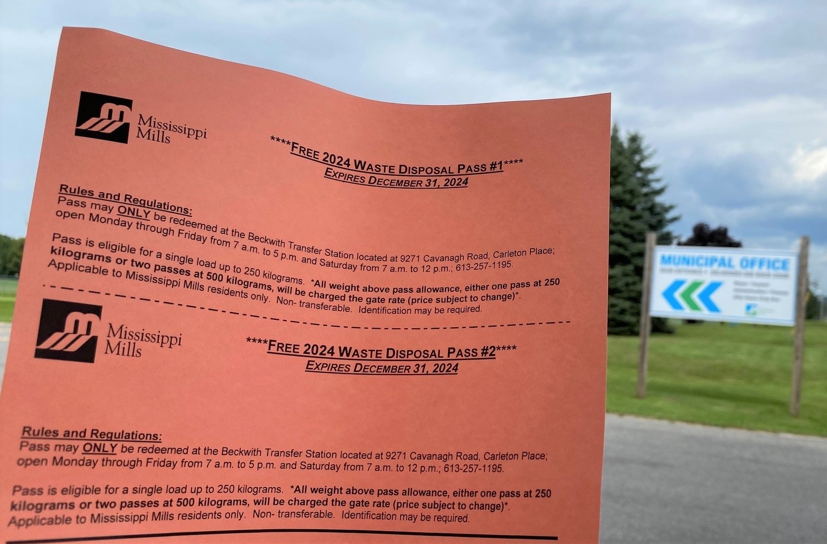 A piece of salmon-coloured paper with the words 'Free 2024 Waste Disposal Pass' is held in front of a large white sign showing the words 'Municipal Office'