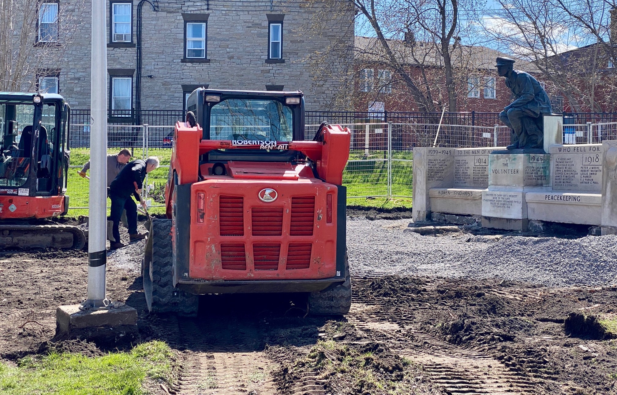 Two men rake dirt in front of a stone Cenotaph