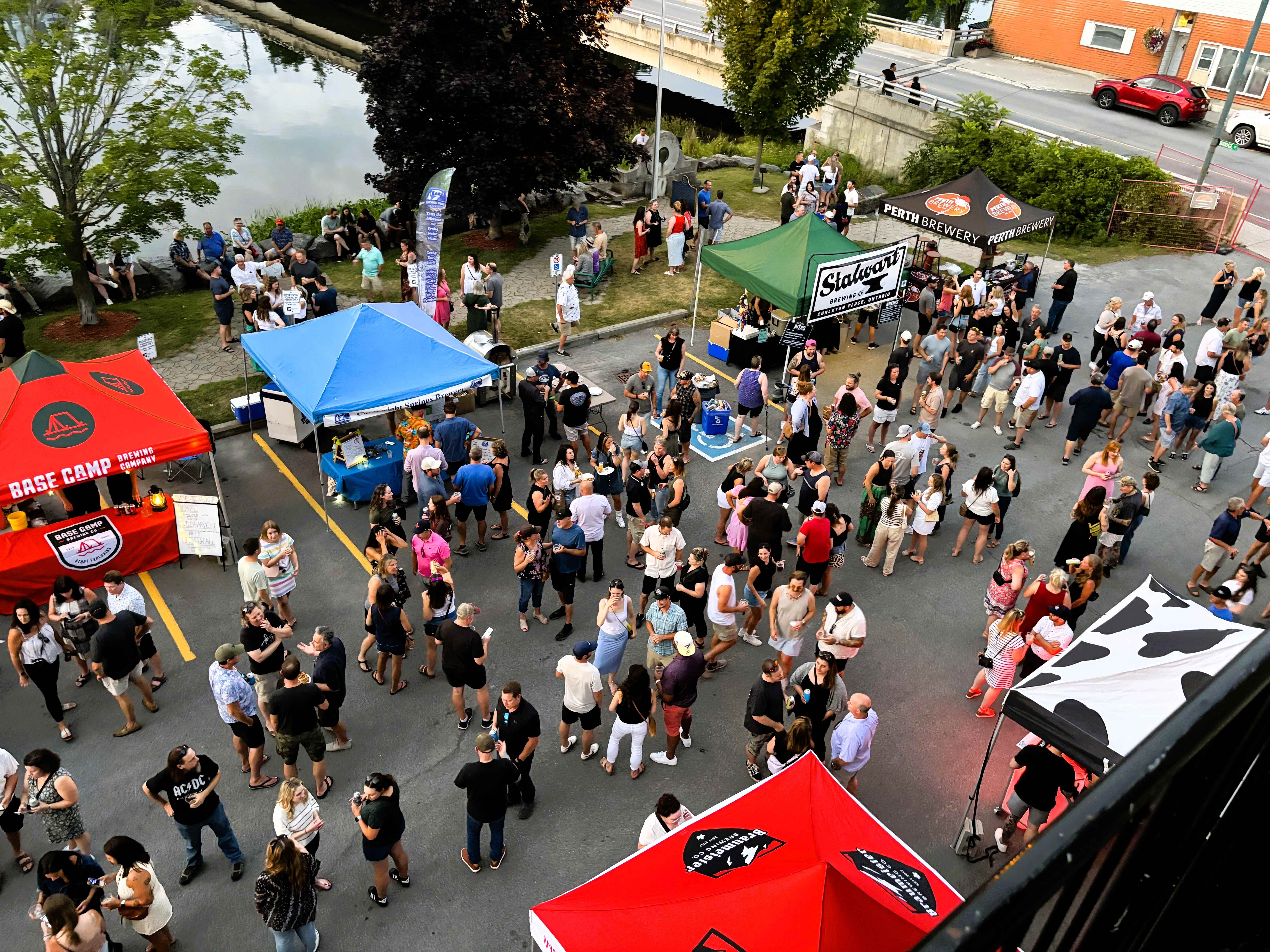 From above, people gather in a parking lot along a river. Tents with vendors are set up.
