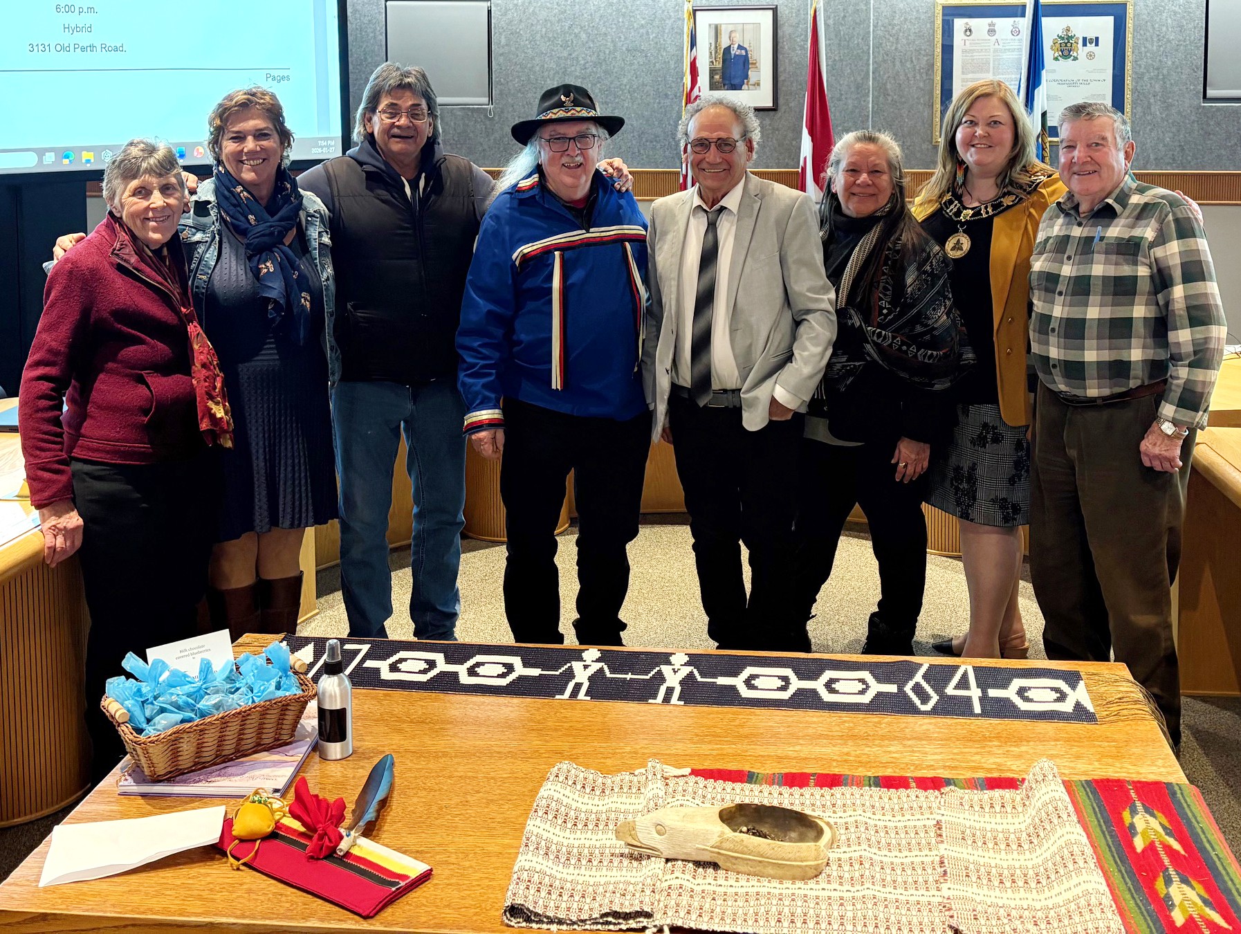 Group of people stand together in Council Chambers for a photo. A table in front shows Indigenous smudging materials and memorabilia.