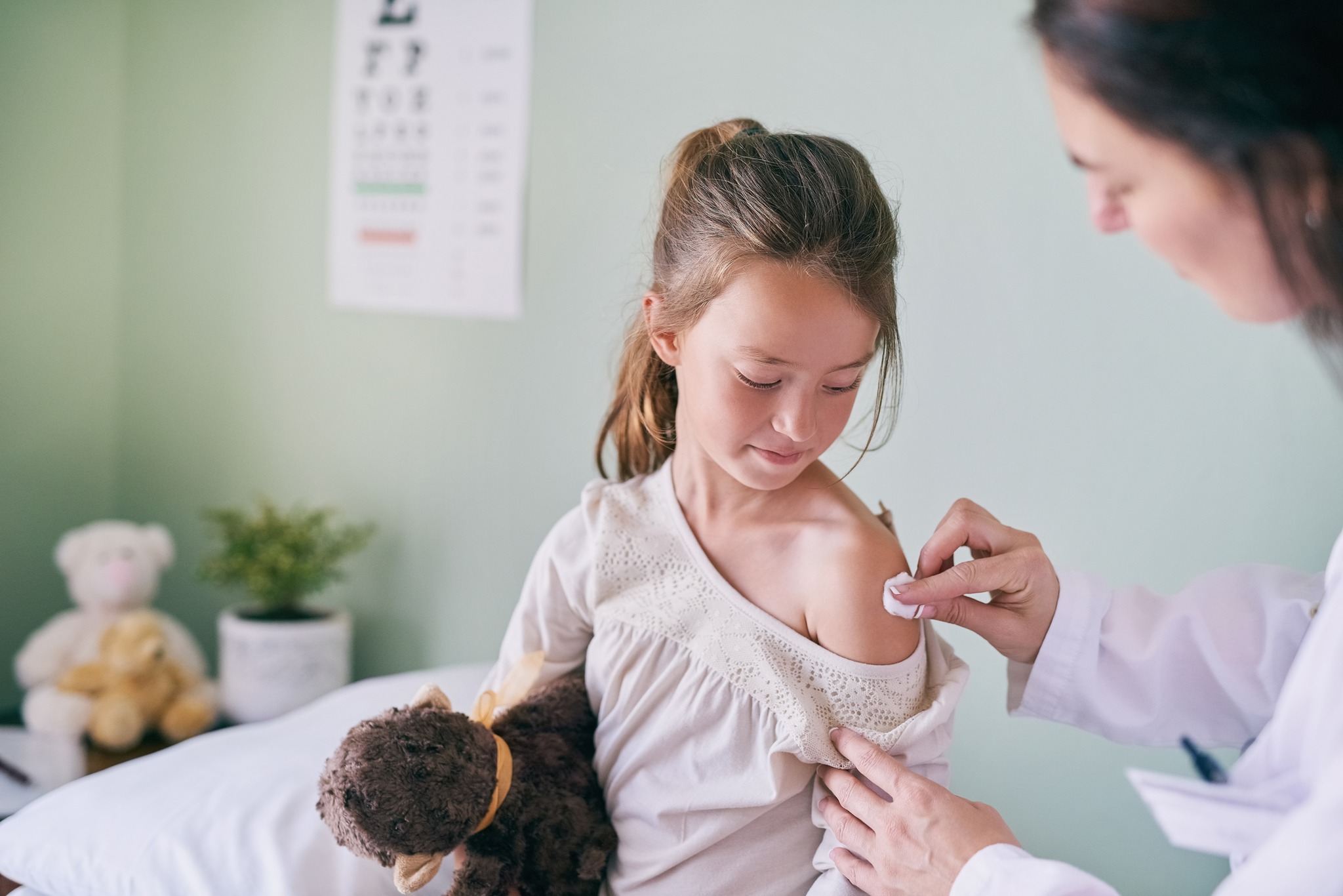 A young girl holds a stuffed animal and sits in a doctor's office while a female doctor gives her a shot