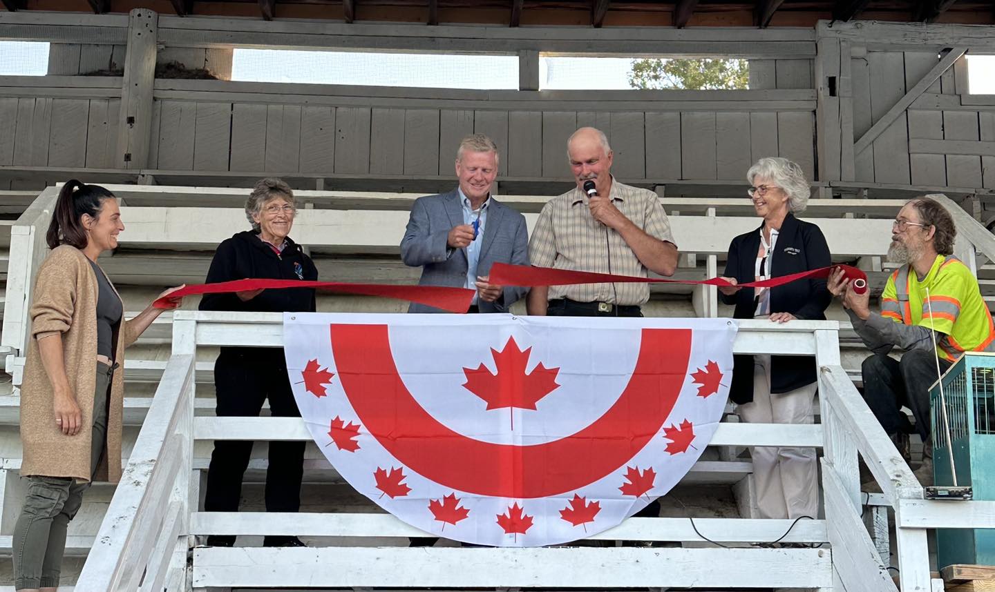 A group of people stand together underneath a covered, white-washed grandstand as a red ribbon is cut.