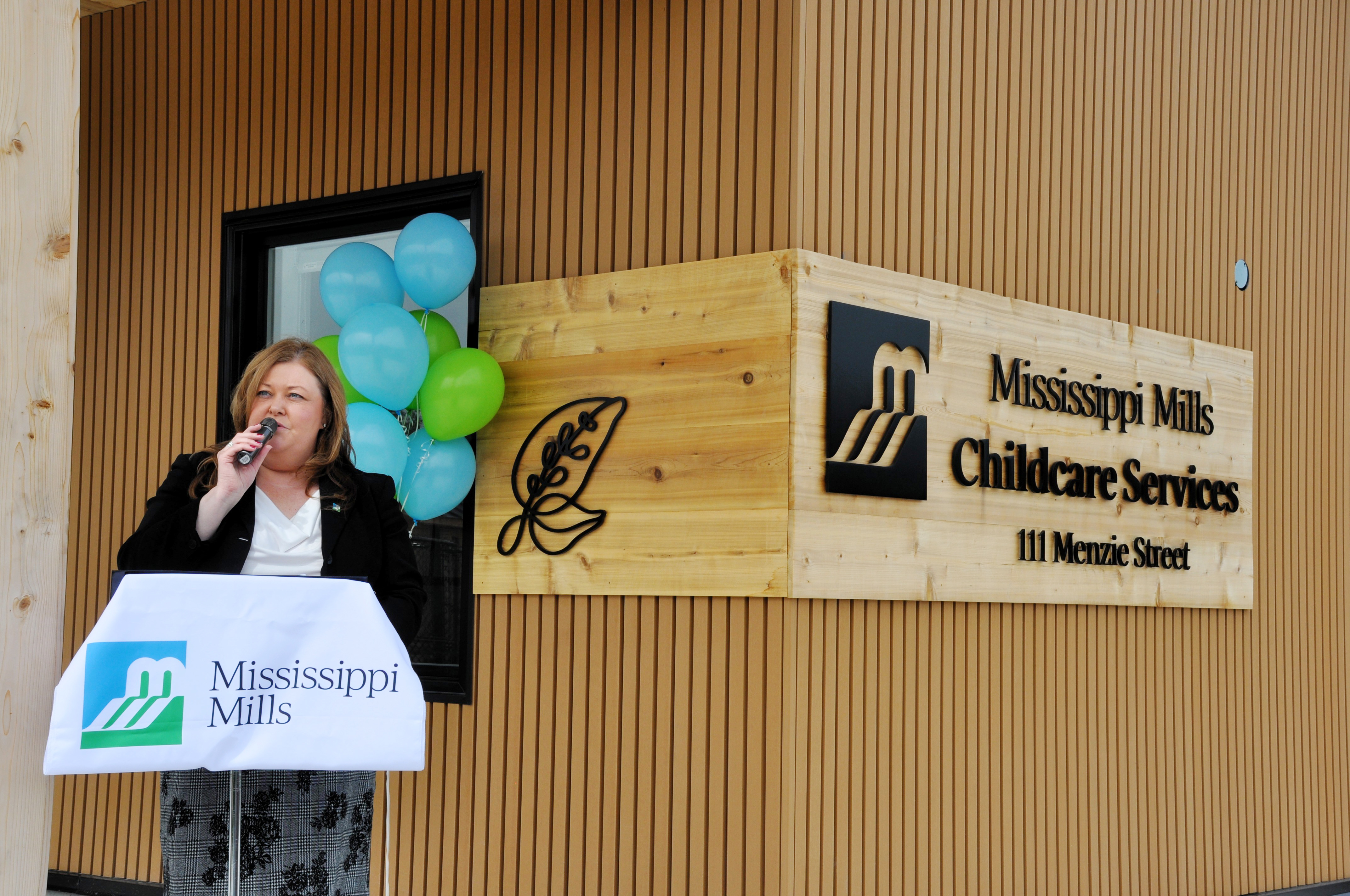 A woman speaks at a lectern with a microphone in her hand. A building behind her has signage reading 'Mississippi Mills Childcare Services.'