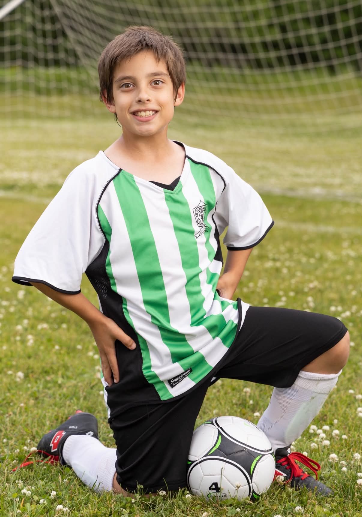 A boy kneels on the soccer field, smiling at the camera, wearing a white and green striped shirt and black shorts. A soccer ball is in front of him.