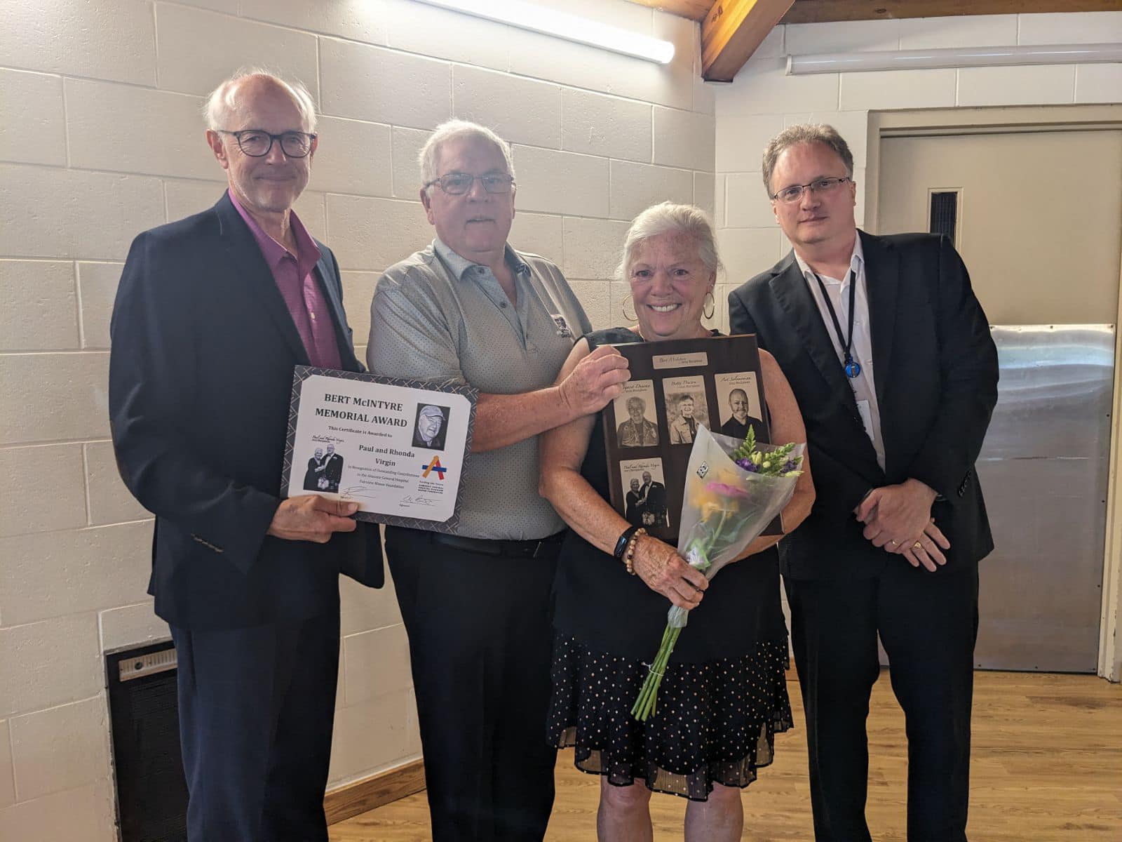 Four people stand together for a photo. The man on the left is holding a certificate, a woman, second from right, holds a photo collage and bouquet of flowers.