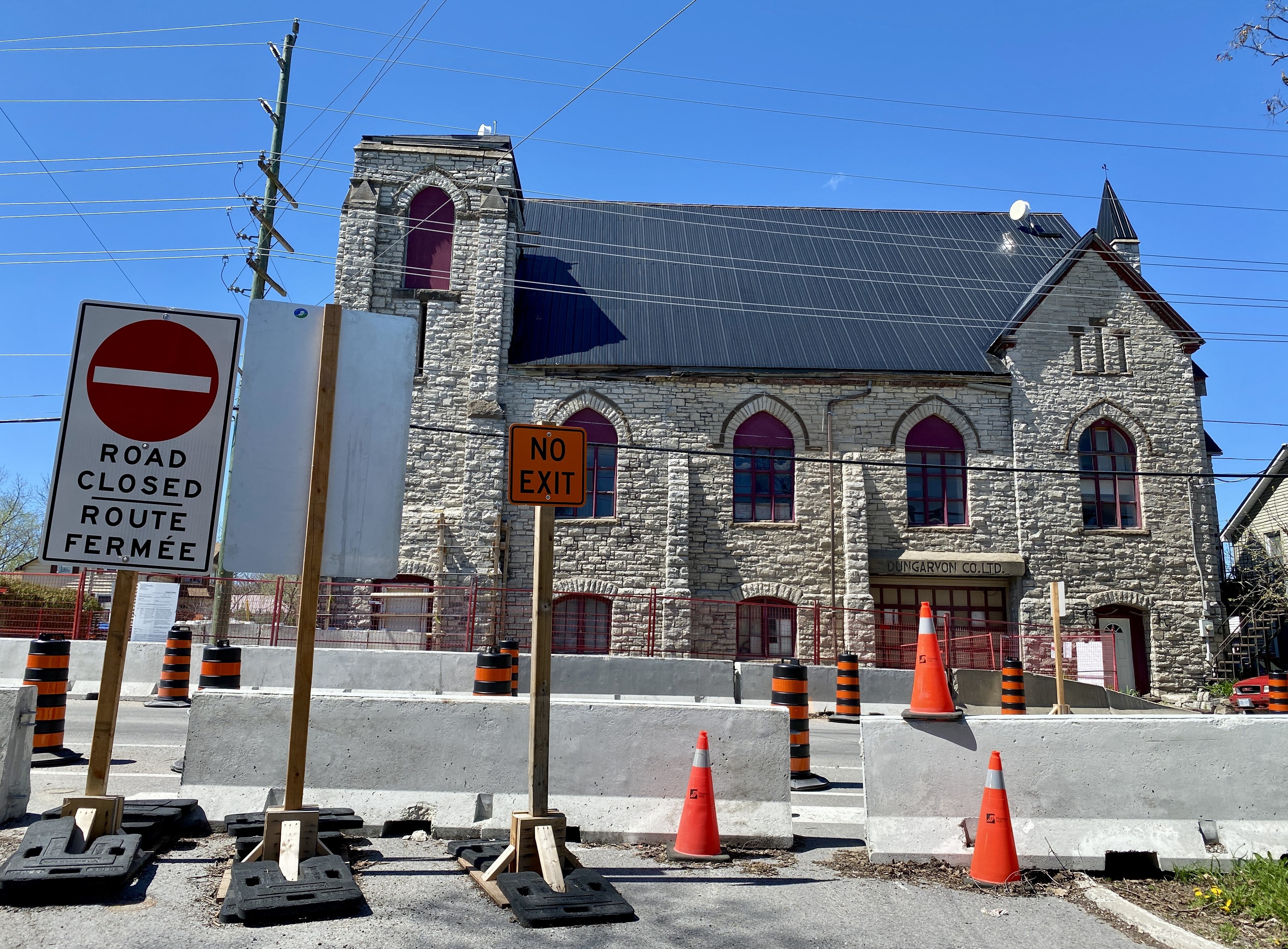 Photo of multi-storey stone building in the background with concrete barriers lined up on a road in front of the building. An orange 'No Exit' sign stands in front with a 'Road Closed' sign next to it.