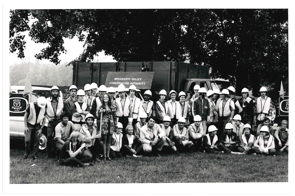 A black and white image of a group of people in light coloured vests and white hard hats gathered for a group photo. A truck and trees are in the background, along with a sign that reads 'Mississippi Valley Conservation Authority.'
