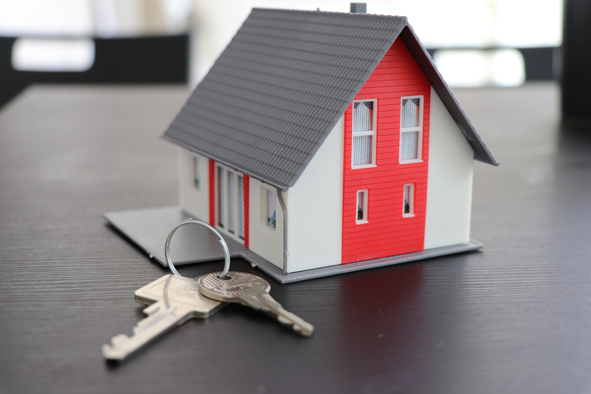 Model of a red and white house with a grey roof sits on a wooden table next to silver house keys
